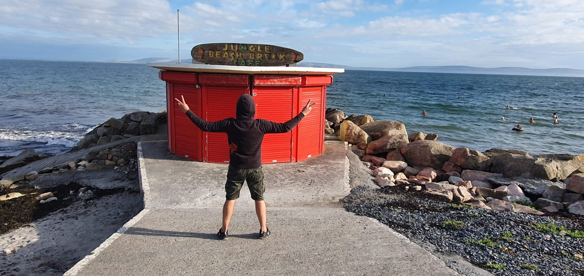 Person standing with arms outstretched facing the ocean at Huntington Beach, near a red beach break stand with a sign that reads "Jungle Beach Break".