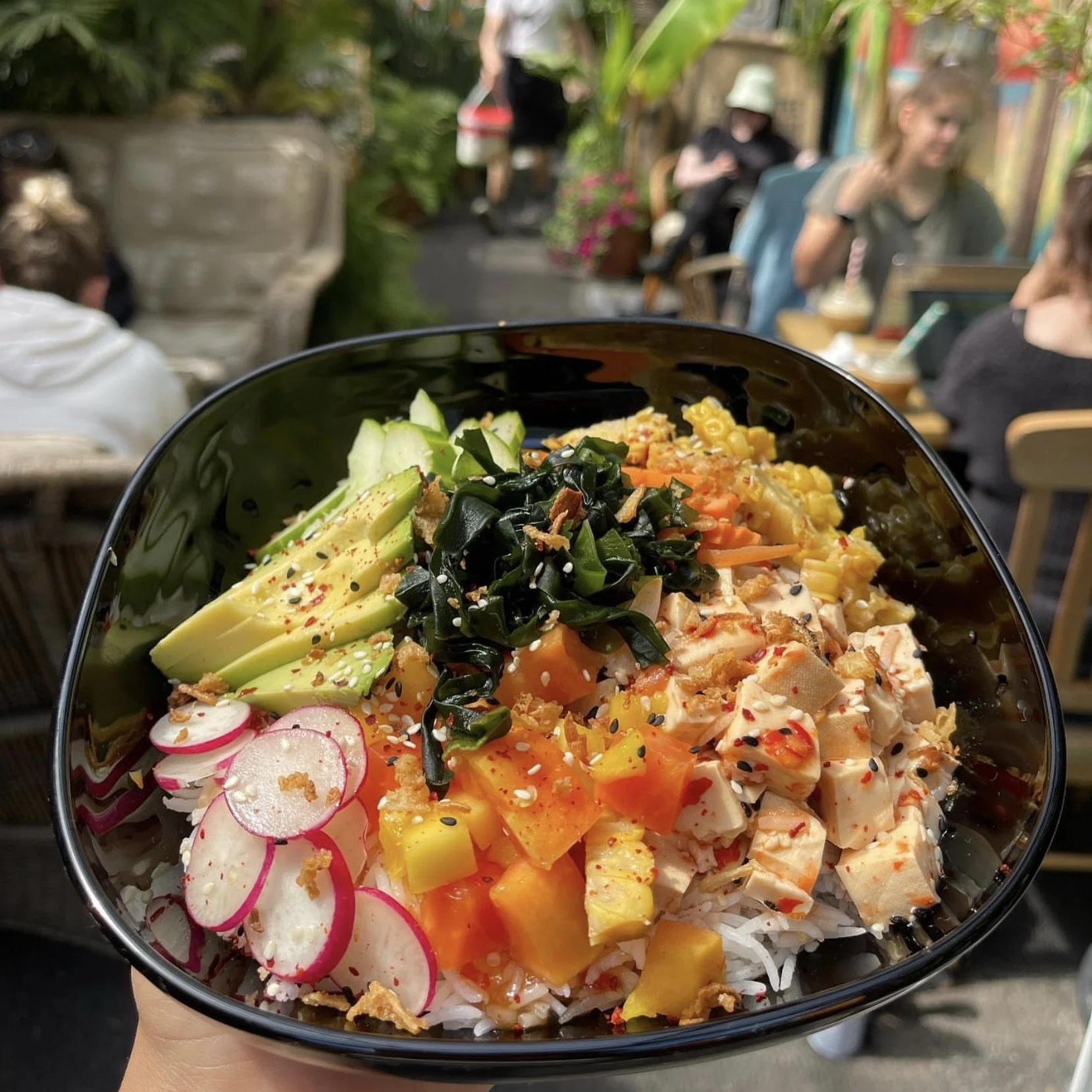 Close-up of a bowl of colorful chopped vegetables, including radishes, cucumbers, seaweed, carrots, corn, and shredded chicken, served with white rice. The background shows a lively outdoor restaurant with people sitting and dining.