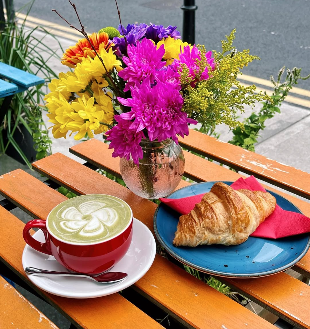 A wooden outdoor table with a vase of colorful flowers, a croissant on a blue plate with a pink napkin, and a cup of matcha latte with latte art.