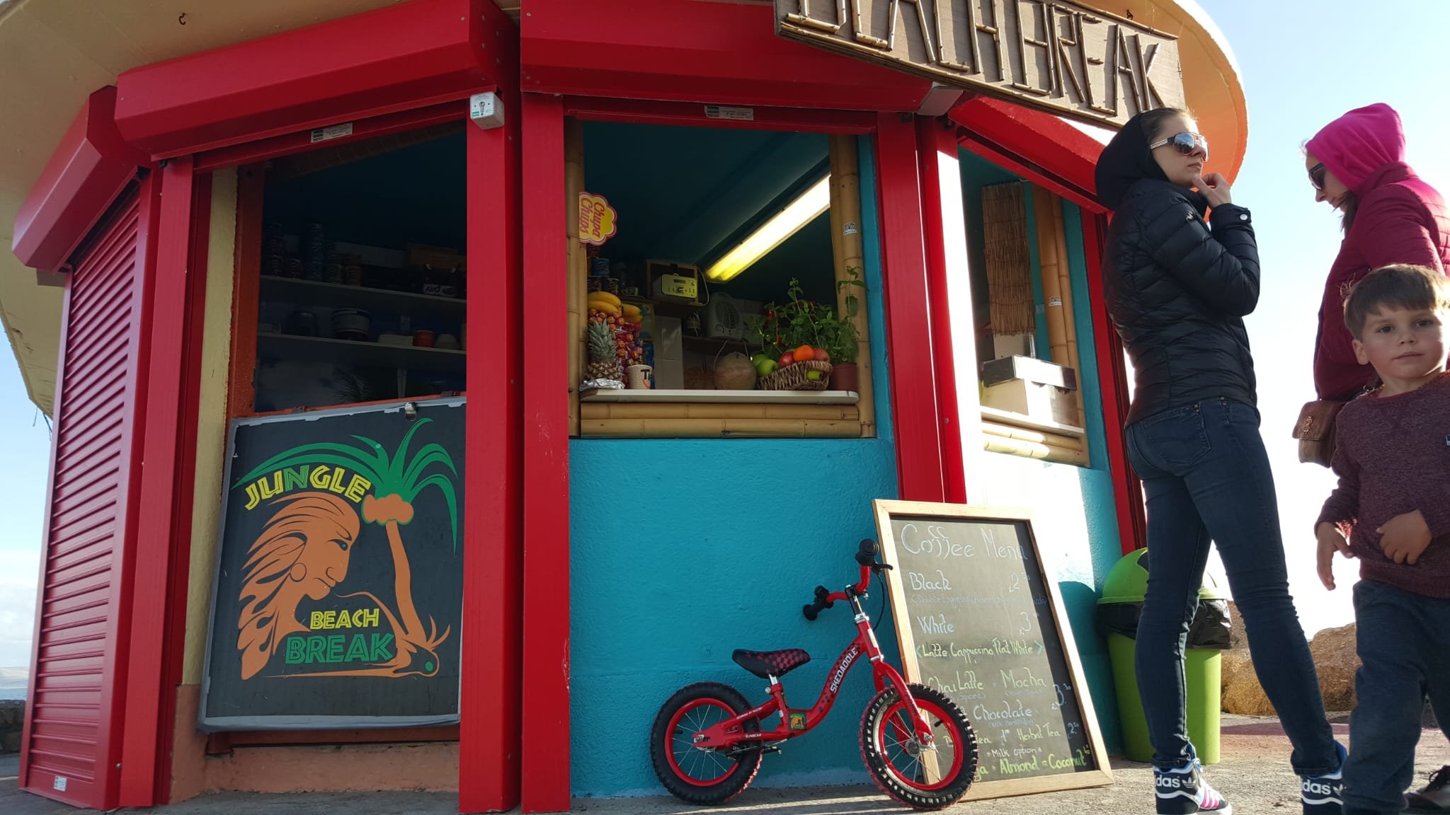 Two women and a young boy standing outside a colorful beachside snack kiosk with a menu board, bicycle, and small fruit stand visible.