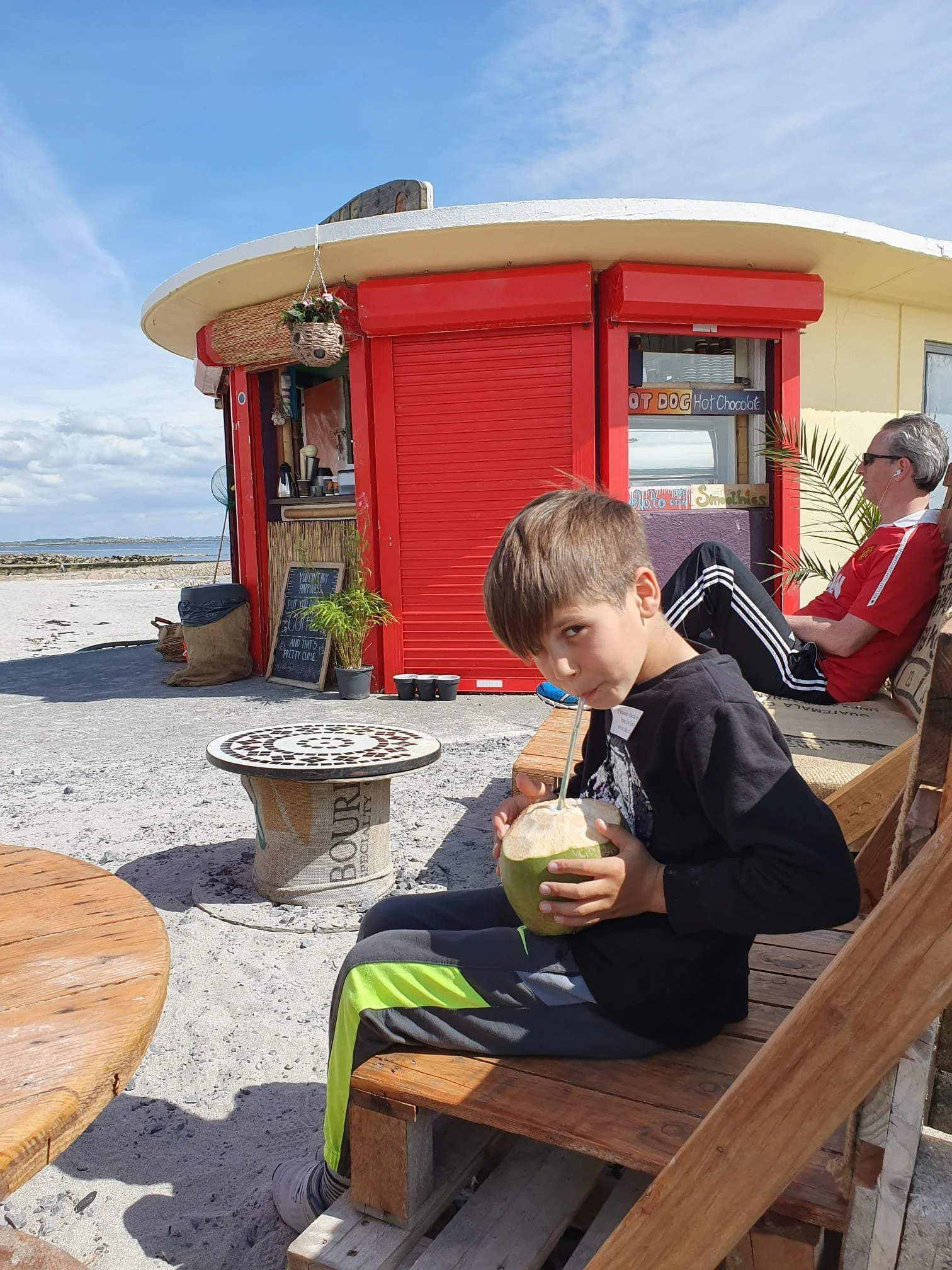 A young boy is sitting on a wooden bench on a beach, drinking from a coconut with a straw. An older man with gray hair and sunglasses is sitting nearby, relaxing in a chair. In the background, there is a red beach shack with a sign offering hot chocolate, smoothies, and other treats, and the sky is partly cloudy.