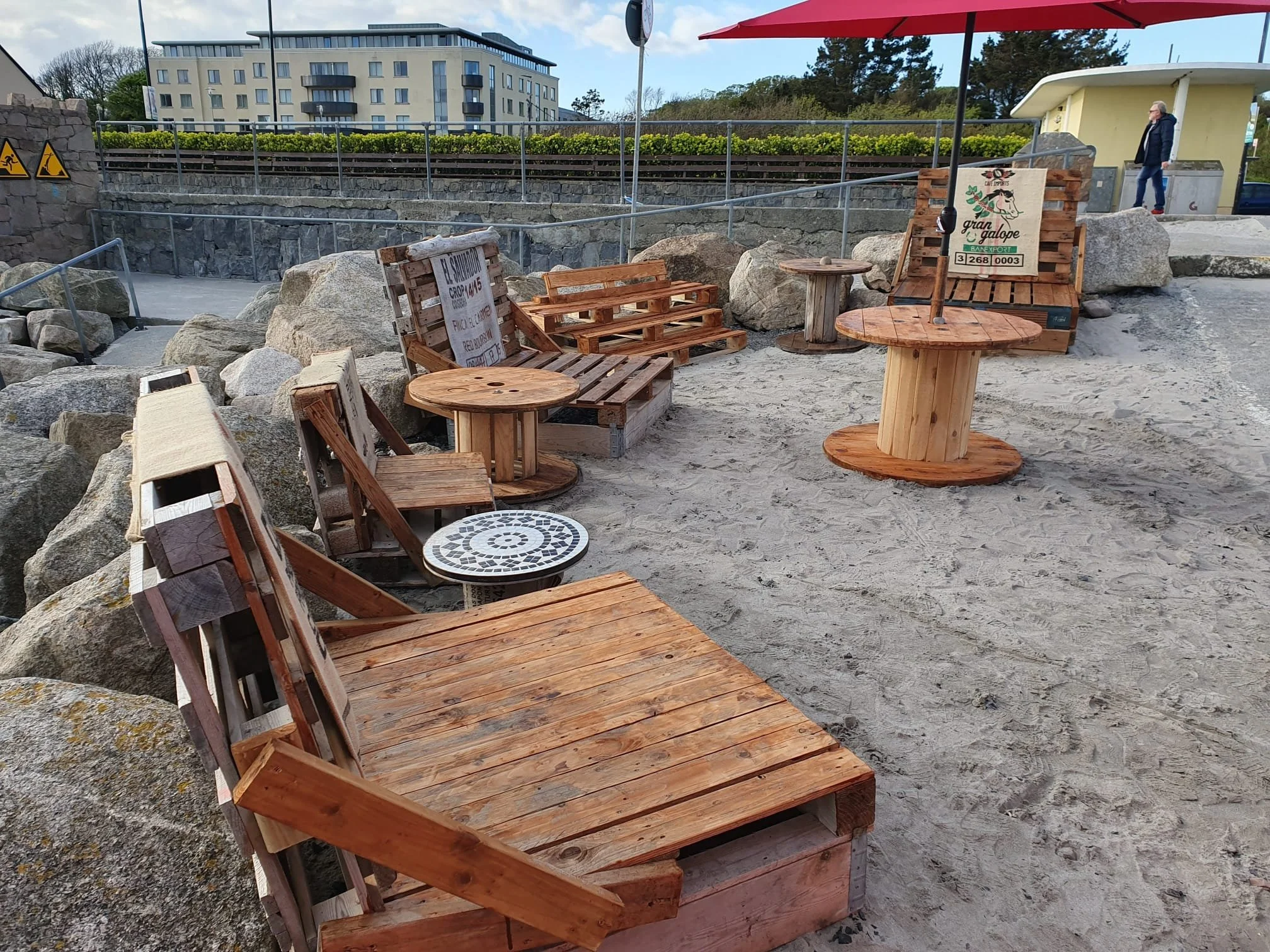 Outdoor seating area with wooden furniture, large rocks, and a sandy surface, with a red umbrella and a sign featuring a horse logo and text. In the background, there are buildings, trees, and a person walking.
