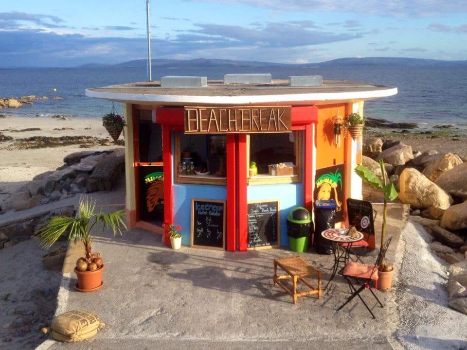 A small colorful beach kiosk with a sign labeled 'Beachbreak' on a sandy beach near the ocean, with rocks and water in the background.