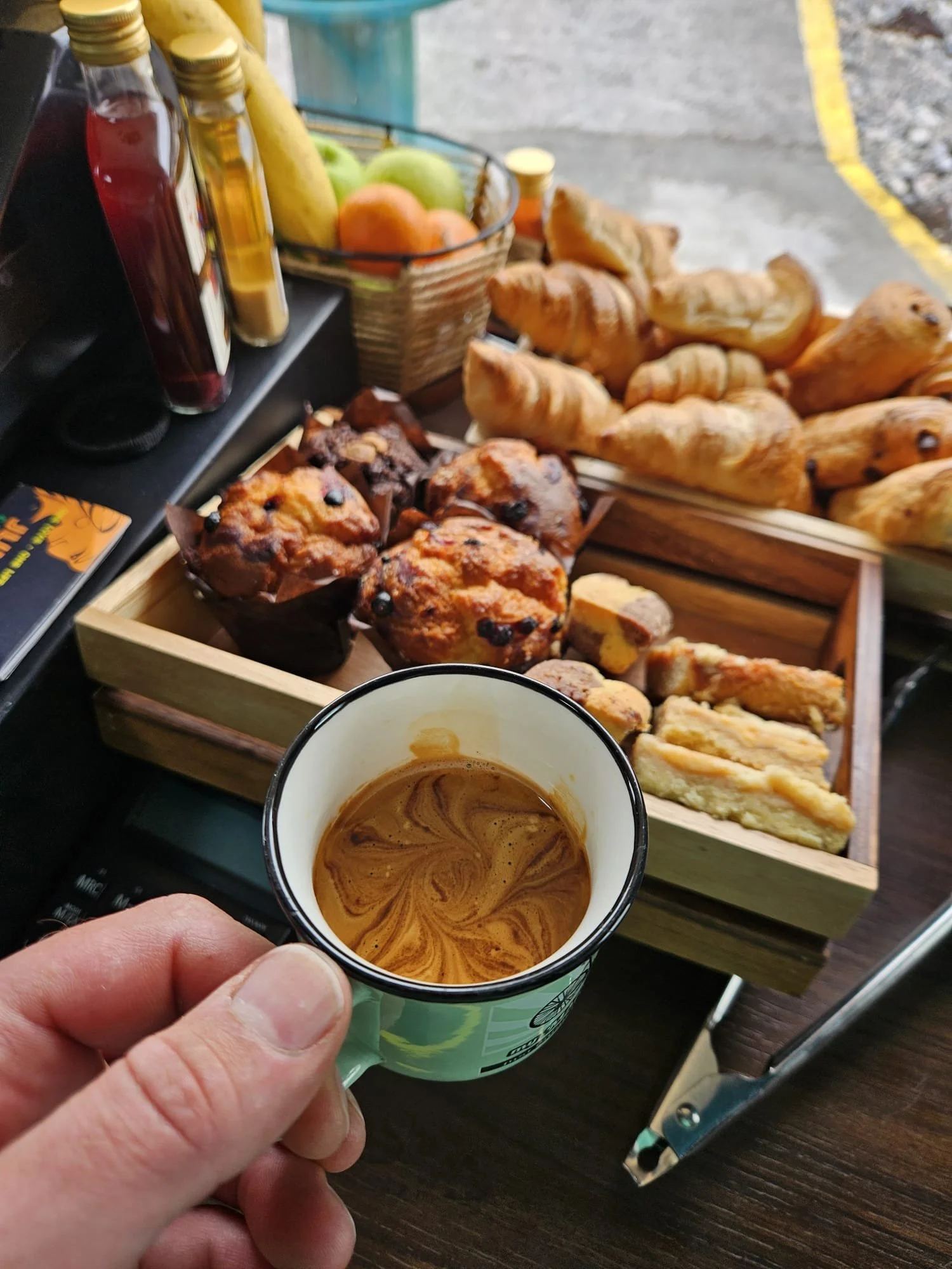Person holding a cup of coffee with a swirl pattern, with a breakfast spread including croissants, muffins, fruit, and bottles of syrup on a table in the background.