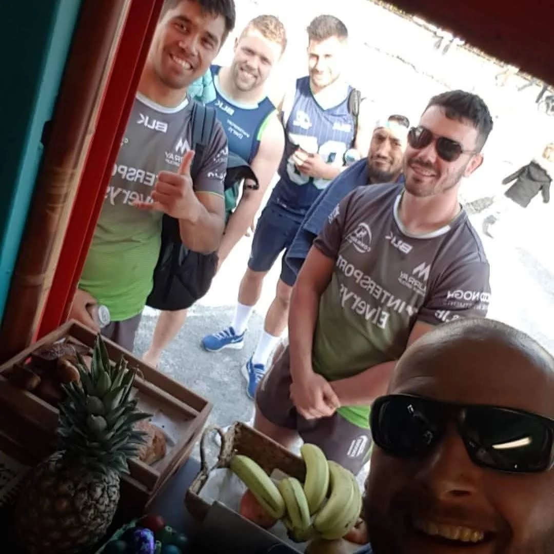 Group of men at a fruit stand, smiling and posing for a photo, with bananas and pineapple in the foreground.