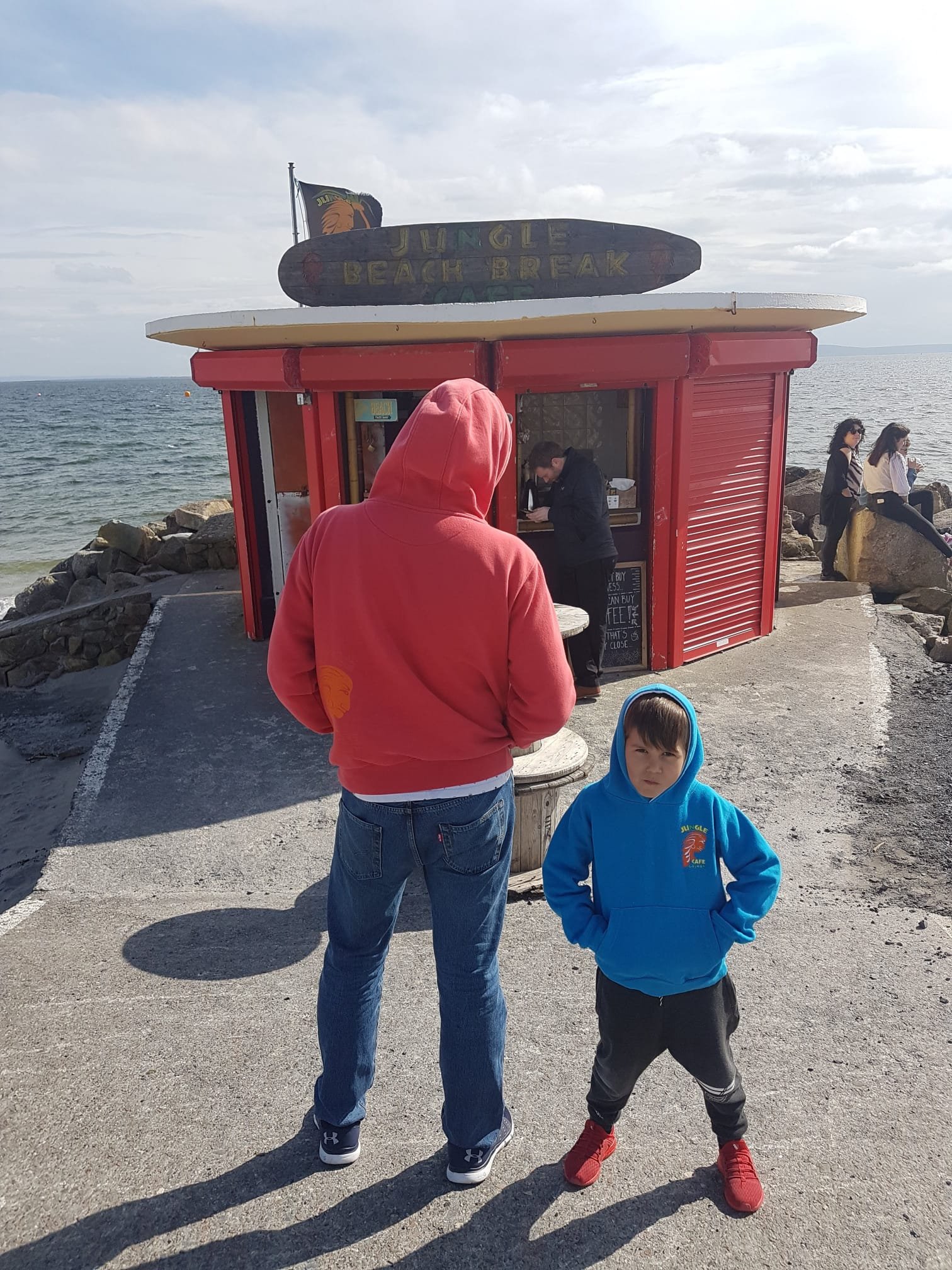 A man in a red hoodie and jeans stands with a young boy in a blue hoodie and black pants near the beach, with a red kiosk behind them. The kiosk has a sign that says 'Jungle Beach Break' and a flag with a lion, and people are sitting on rocks by the shore.
