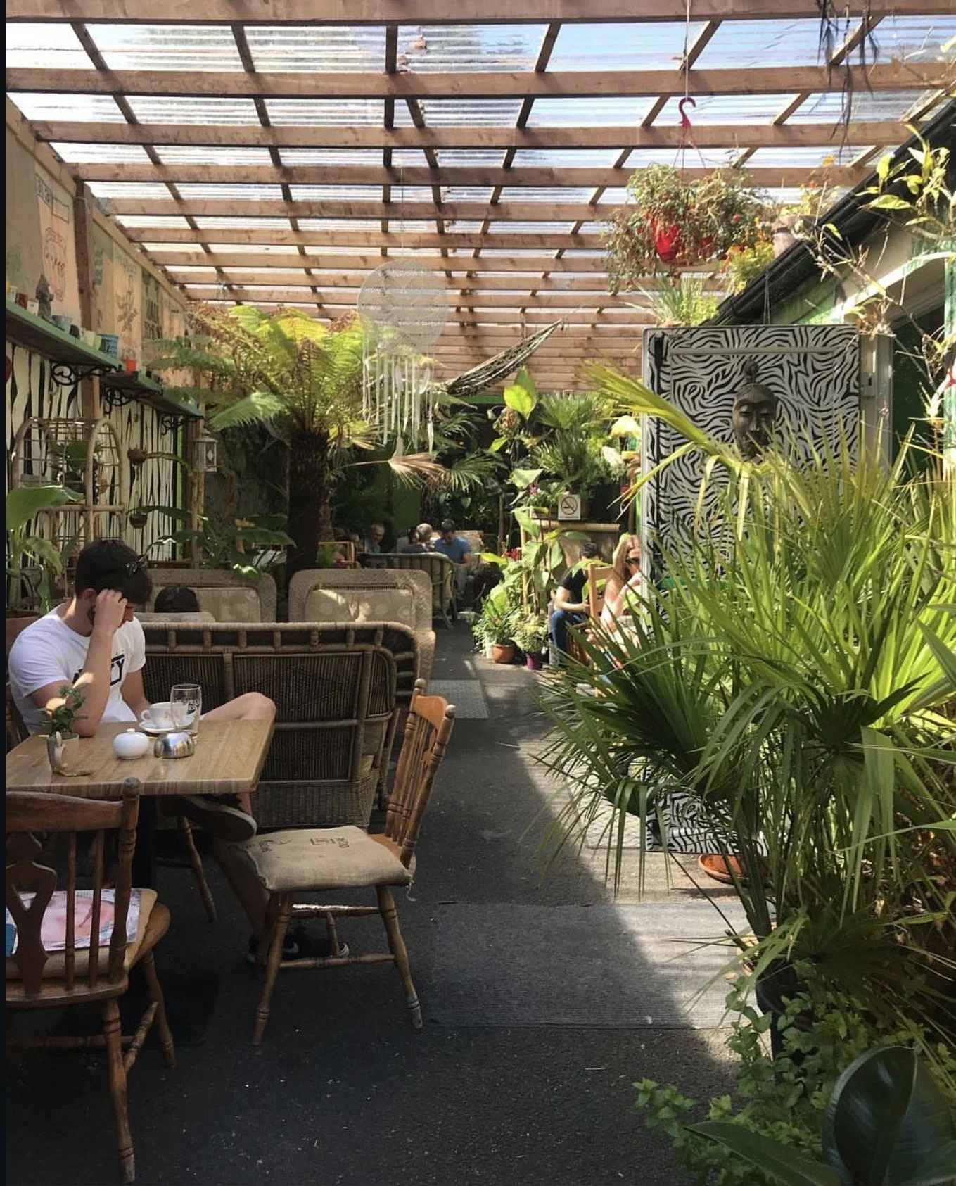 A covered outdoor dining area with wooden and glass roofing, lush green plants, and several people seated at tables enjoying food and drinks.