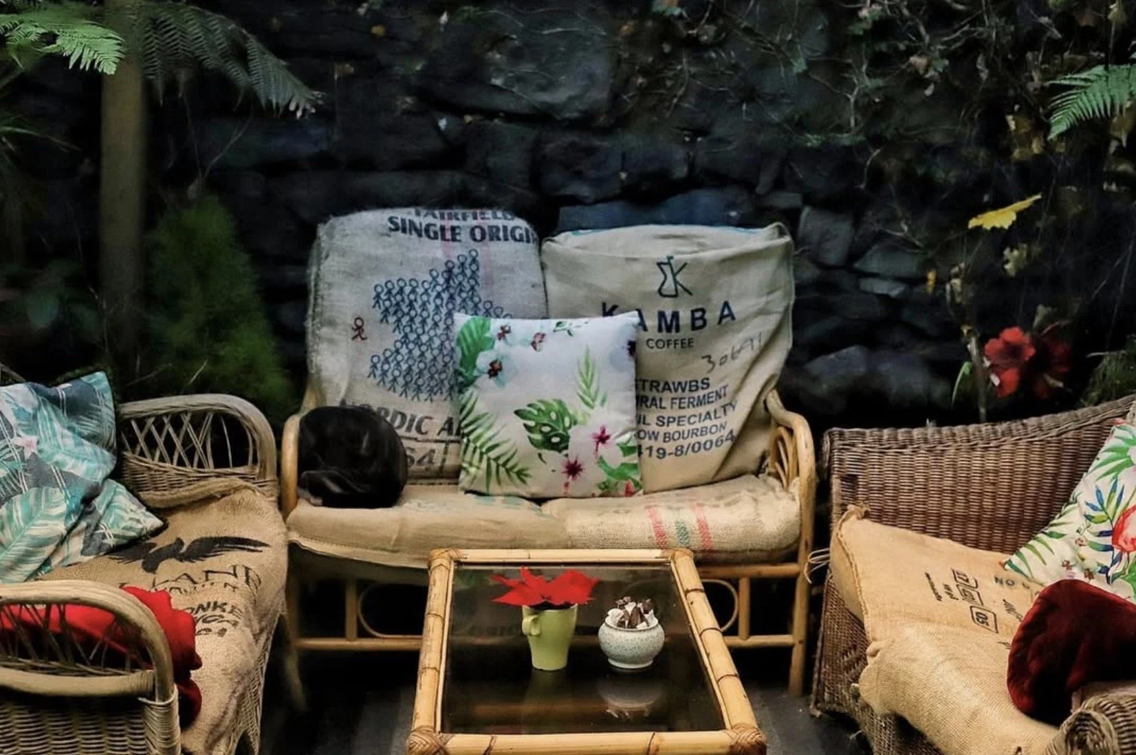Outdoor seating area with wicker and rattan chairs and sofas, decorated with colorful cushions and throw pillows, with a small bamboo table holding a green vase with a red flower and decorative object. The background has a dark stone wall with plants and flowers.