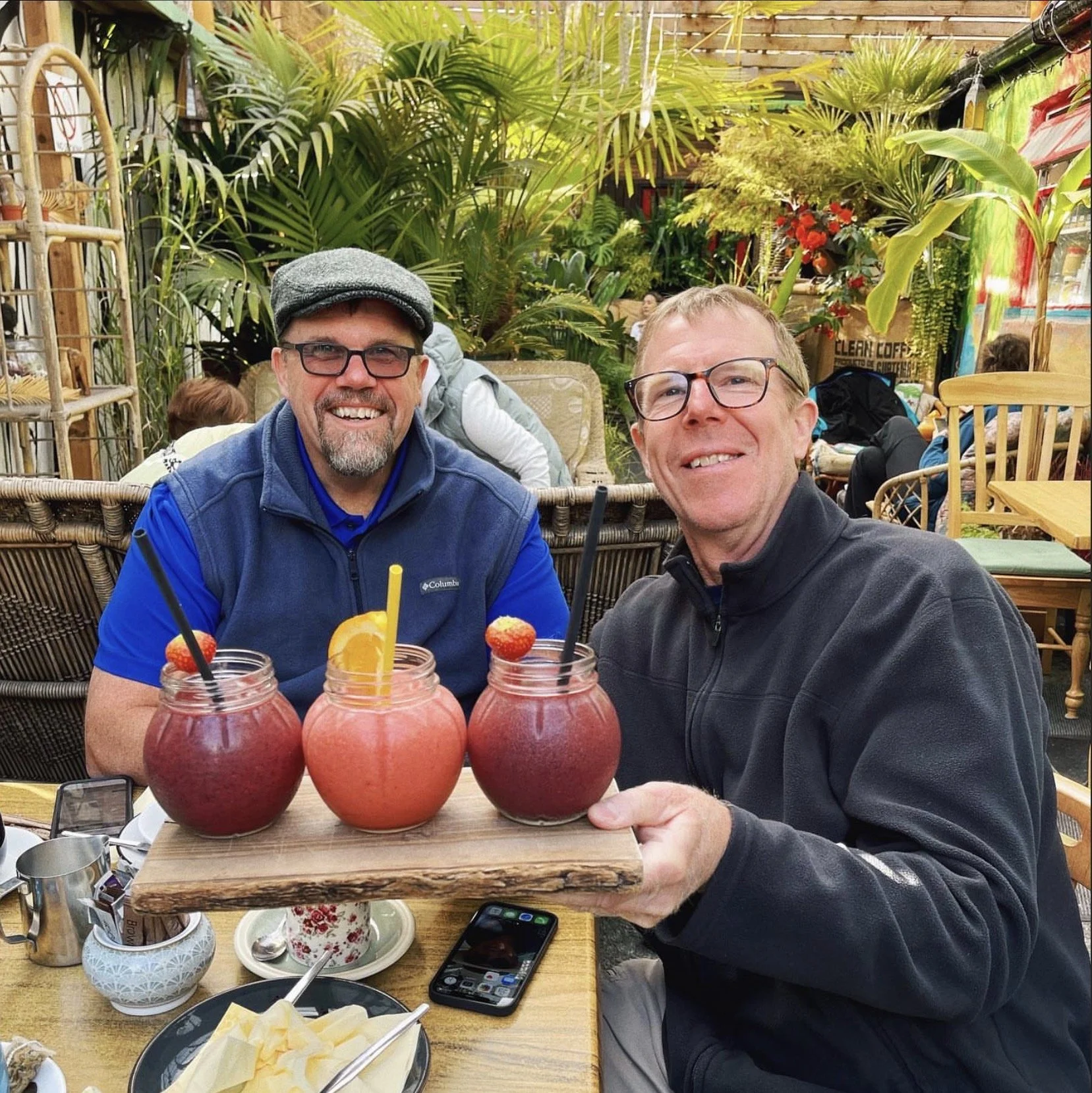 Two men smiling and sitting at a table in a lush indoor garden cafe, holding a tray with three pink smoothies garnished with strawberries and orange slices, with plates of pineapple and ice nearby.