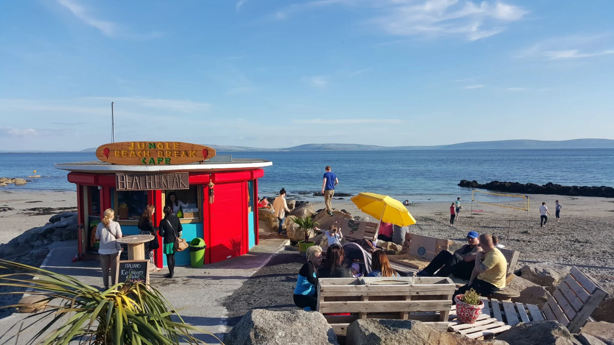 Beach scene with a red and blue beachfront cafe labeled 'Jungle Beach Break Cafe', with people sitting and standing nearby, a yellow umbrella, volleyball game, rocky shoreline, and calm ocean with distant hills under a partly cloudy sky.