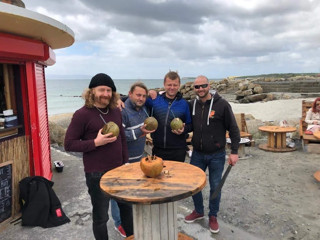 Four men standing outdoors on a beach holding coconuts, with a woman sitting on a bench in the background, overcast sky, standing next to a red food stand, with a round wooden table and rocks in the background.