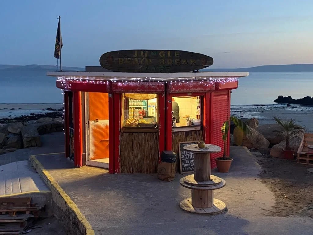 A small beachside food stand with a surfboard sign on top, decorated with string lights, located near the ocean at dusk, with a table, potted plant, and seaside rocks.
