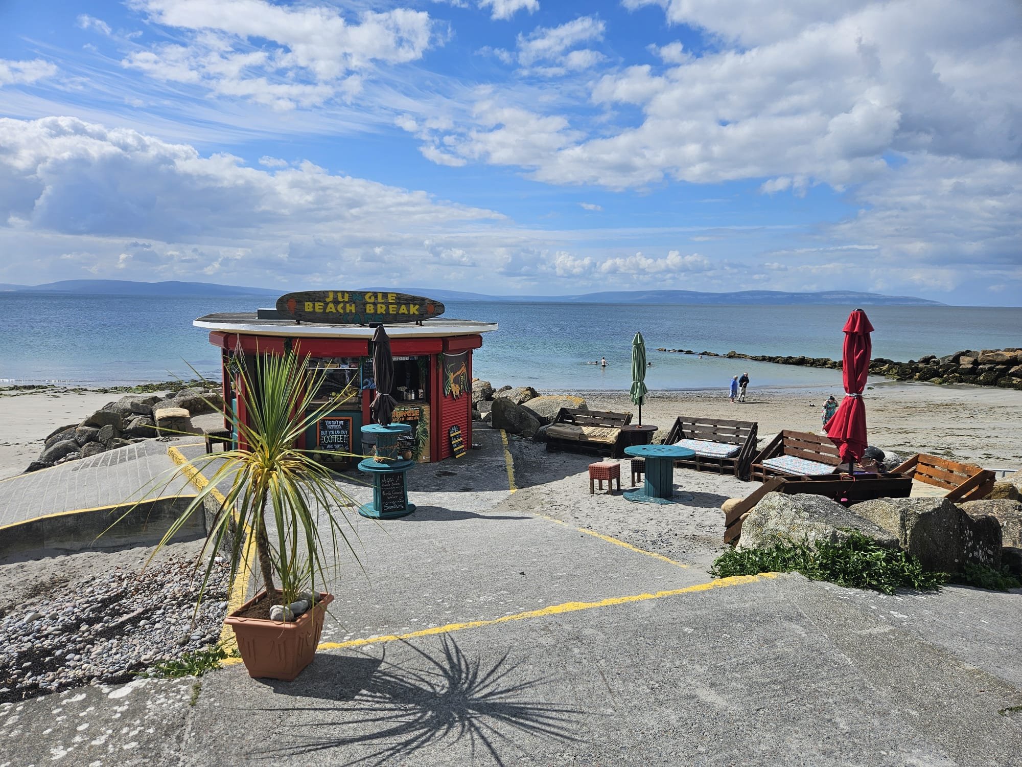A beach scene with a small colorful snack stand labeled 'Jungle Beach Break,' surrounded by benches, umbrellas, and potted plants. The ocean with a few people swimming and a partly cloudy sky are visible in the background.