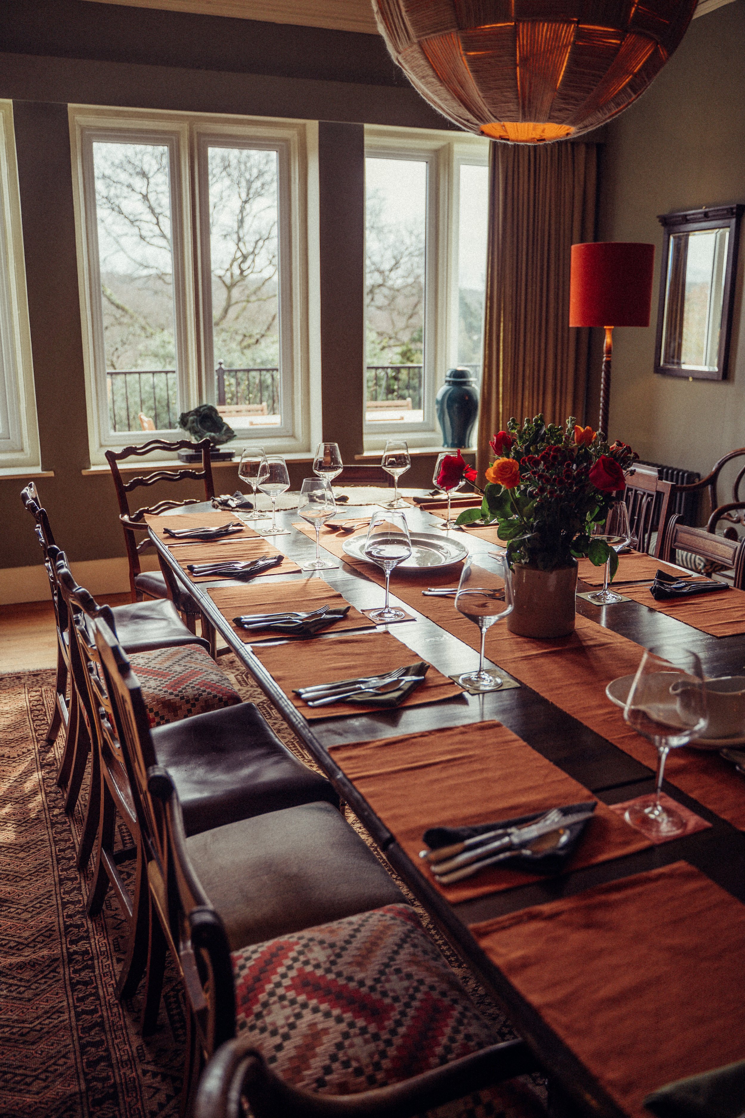 A dining room with a long wooden table set for a meal, featuring wine glasses, napkins, and a floral centerpiece, with large windows in the background.