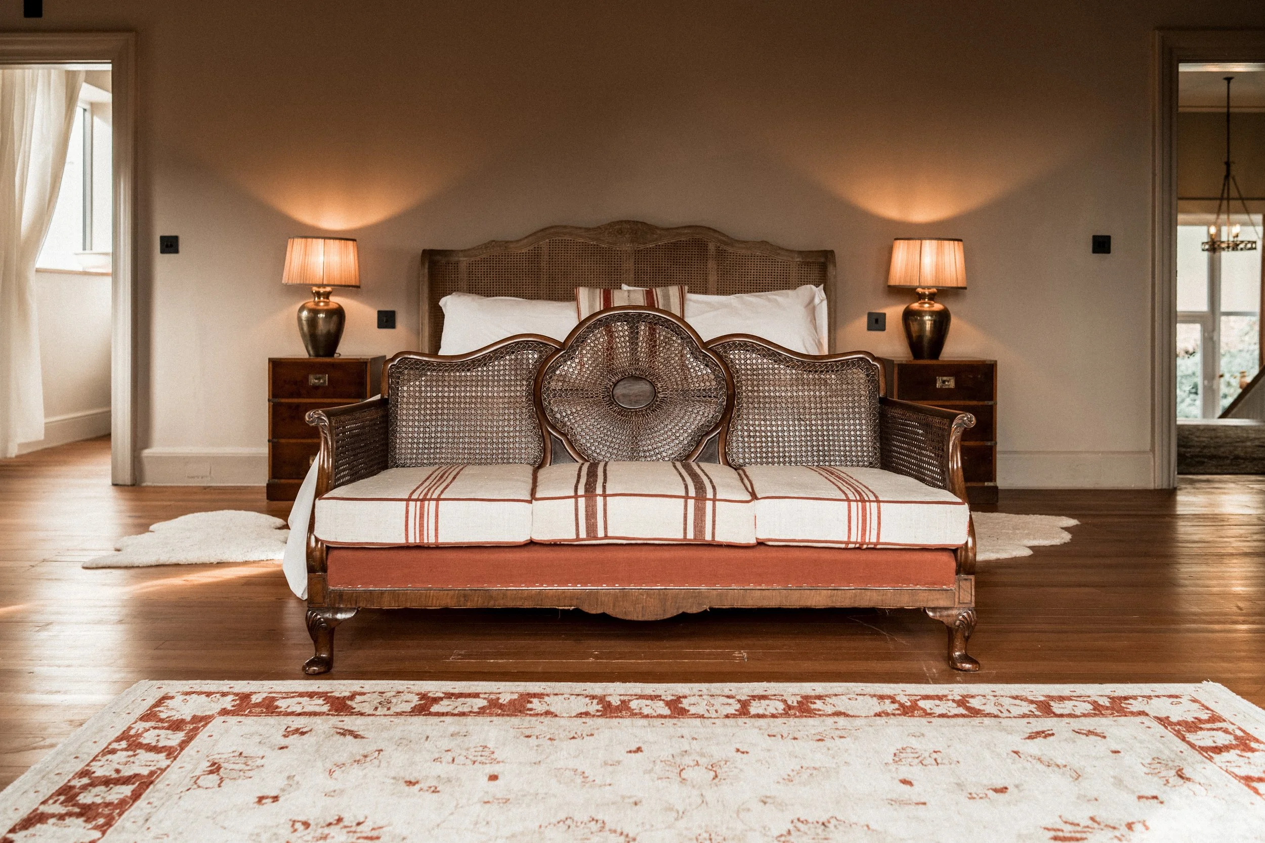 A vintage-style bedroom with a wooden bed and matching sofa featuring woven cane details, two nightstands with lamps, and a large area rug.