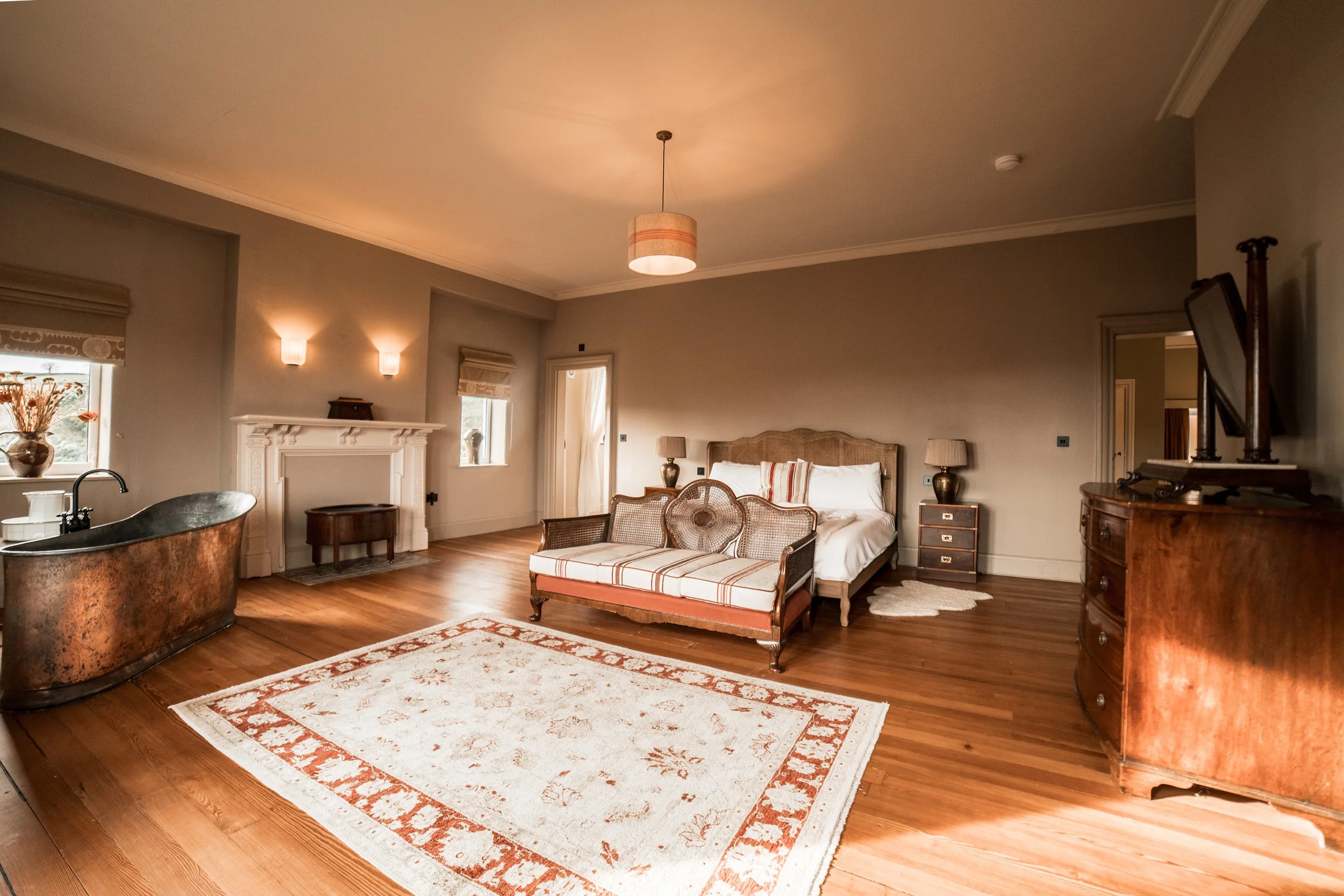 A spacious bedroom with hardwood floors, a large bed with a headboard, two bedside lamps, a vintage-style bench at the foot of the bed, a rug, a fireplace, a copper bathtub, and a dresser with a flat-screen TV.