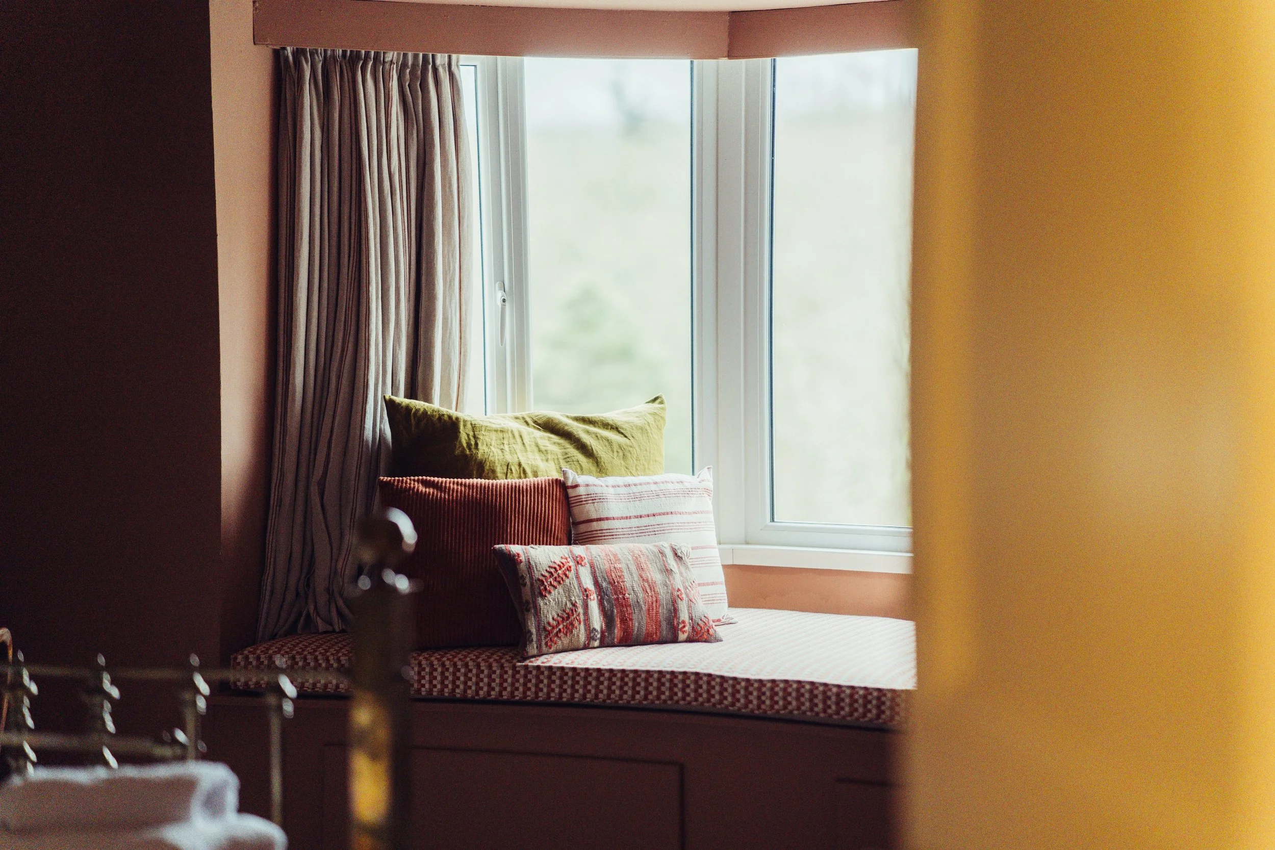 A cozy window nook with a cushioned bench next to a large window, decorated with colorful pillows and beige curtains.