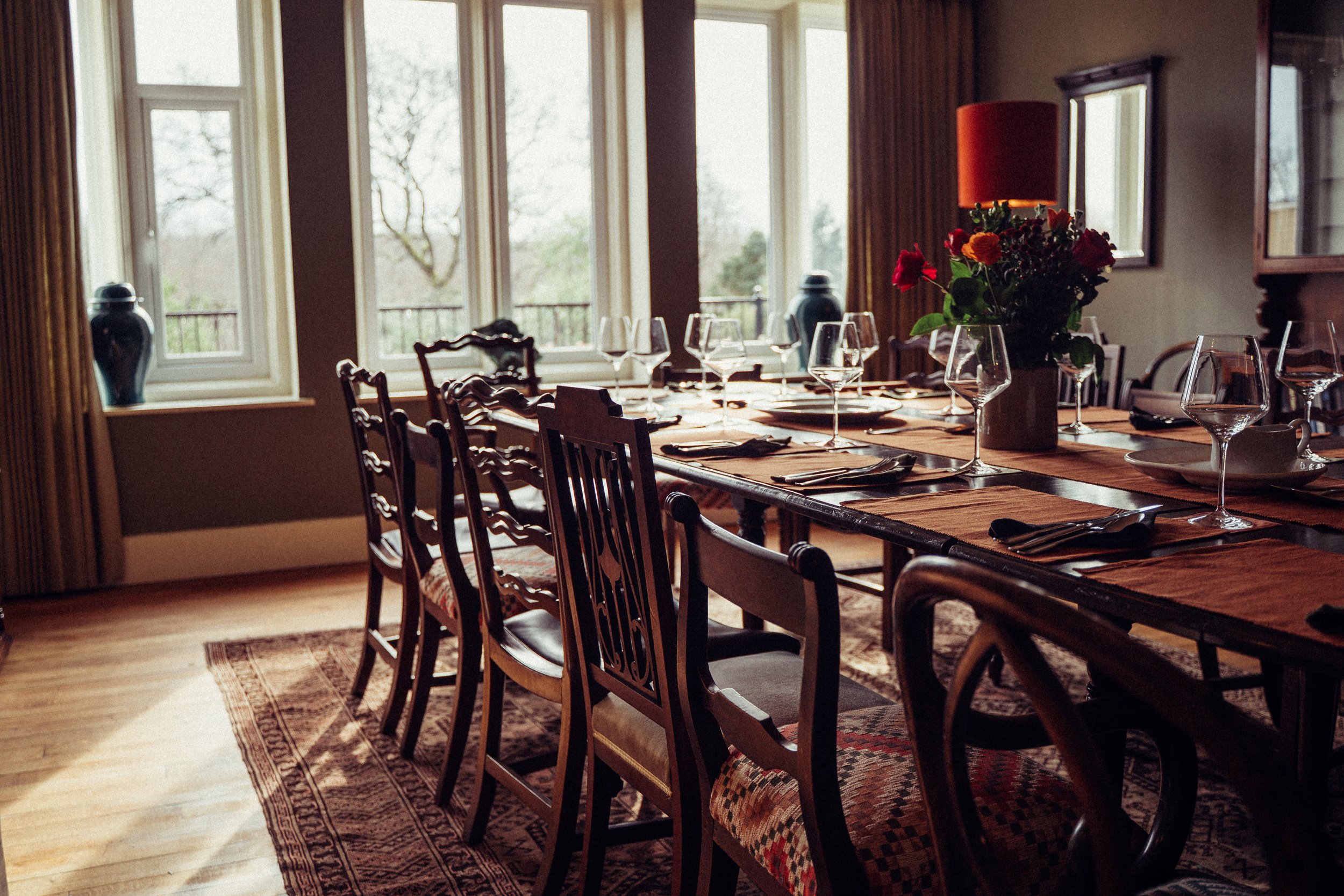 A dining room with a long wooden table, set for a meal with wine glasses, cutlery, and napkins, and decorated with a vase of red and orange flowers. The room has large windows overlooking a garden, with a patterned rug on the floor and framed art on the walls.