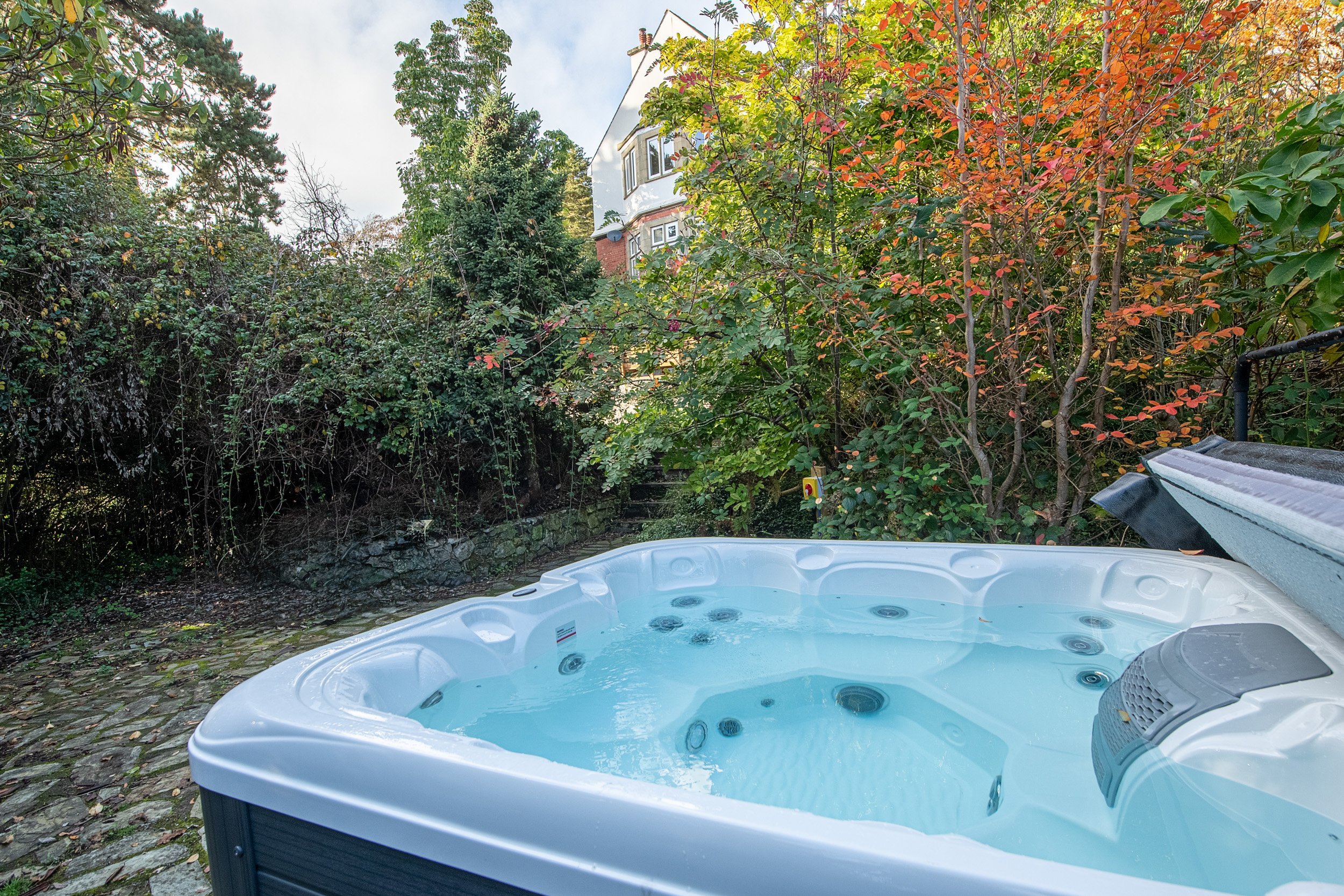 A hot tub filled with water is situated outdoors on a stone-paved surface, surrounded by lush green and red foliage, with a white house visible in the background.