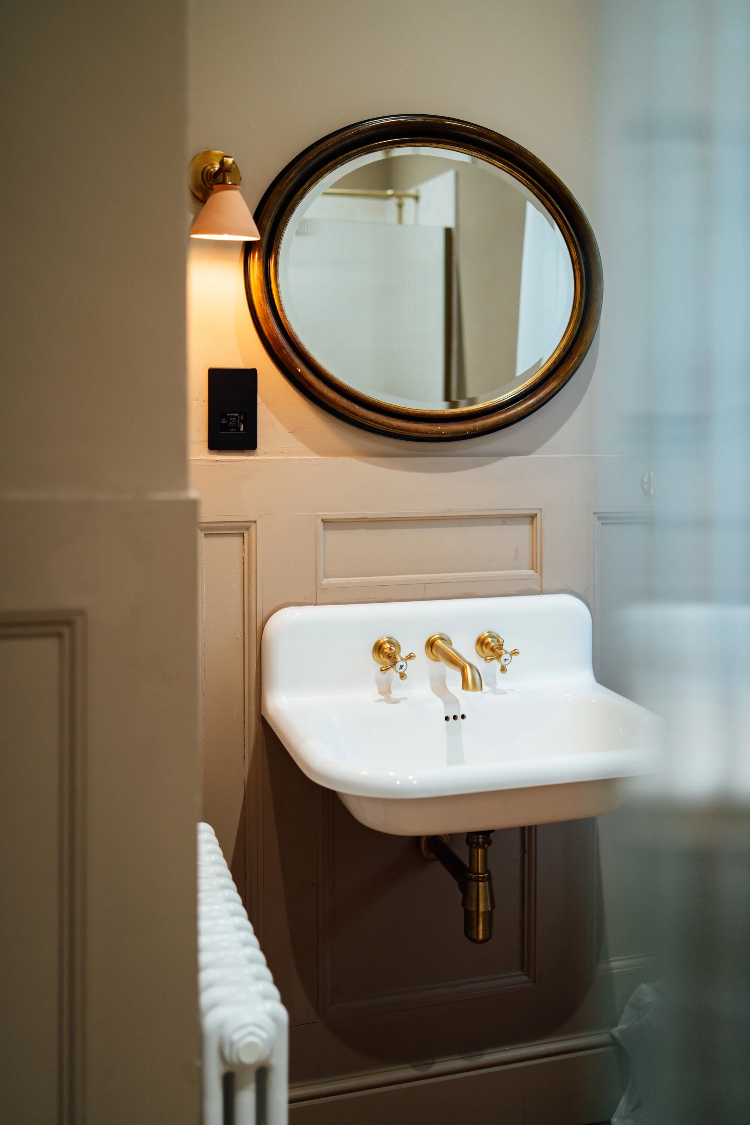 A vintage-style bathroom with a white wall-mounted sink featuring gold fixtures, a round mirror with a gold frame, a wall-mounted light, and a radiator.