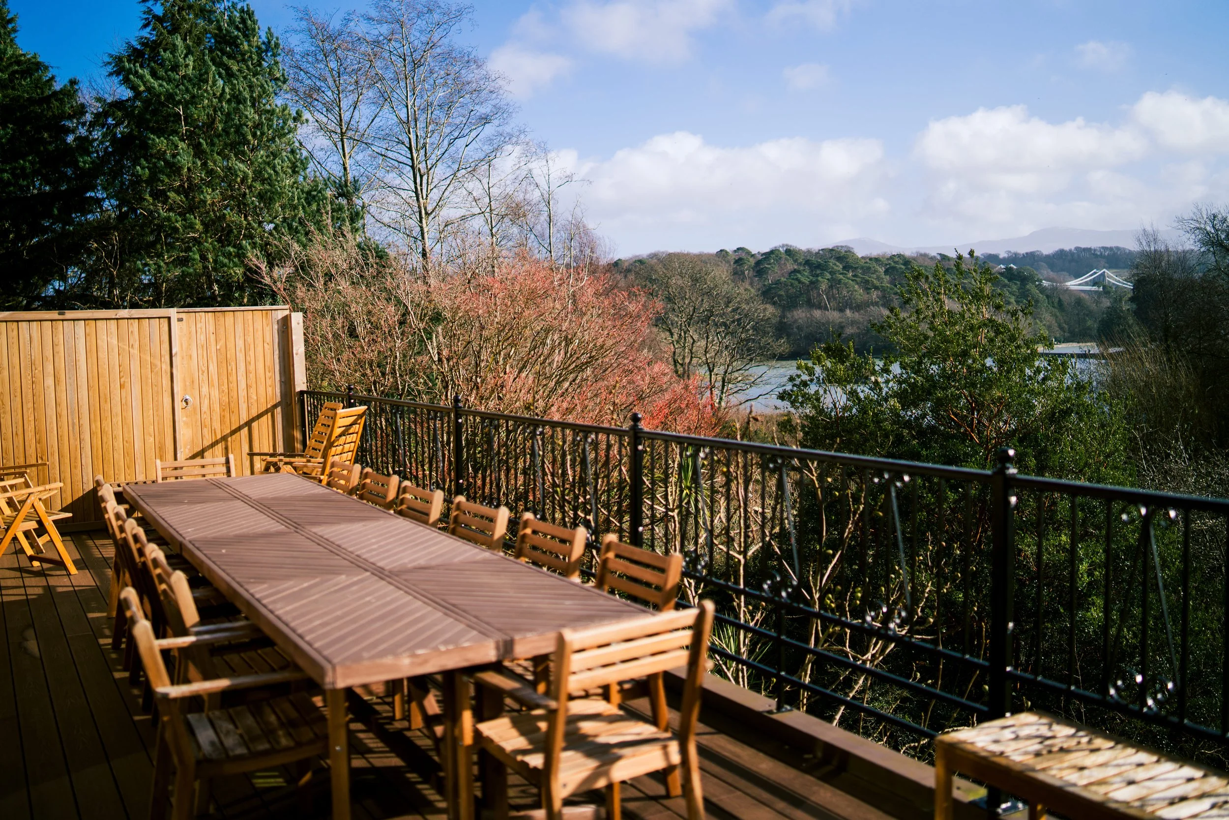 An outdoor wooden deck with a long table and chairs, overlooking a scenic view of trees, a river, and mountains under a partly cloudy sky.
