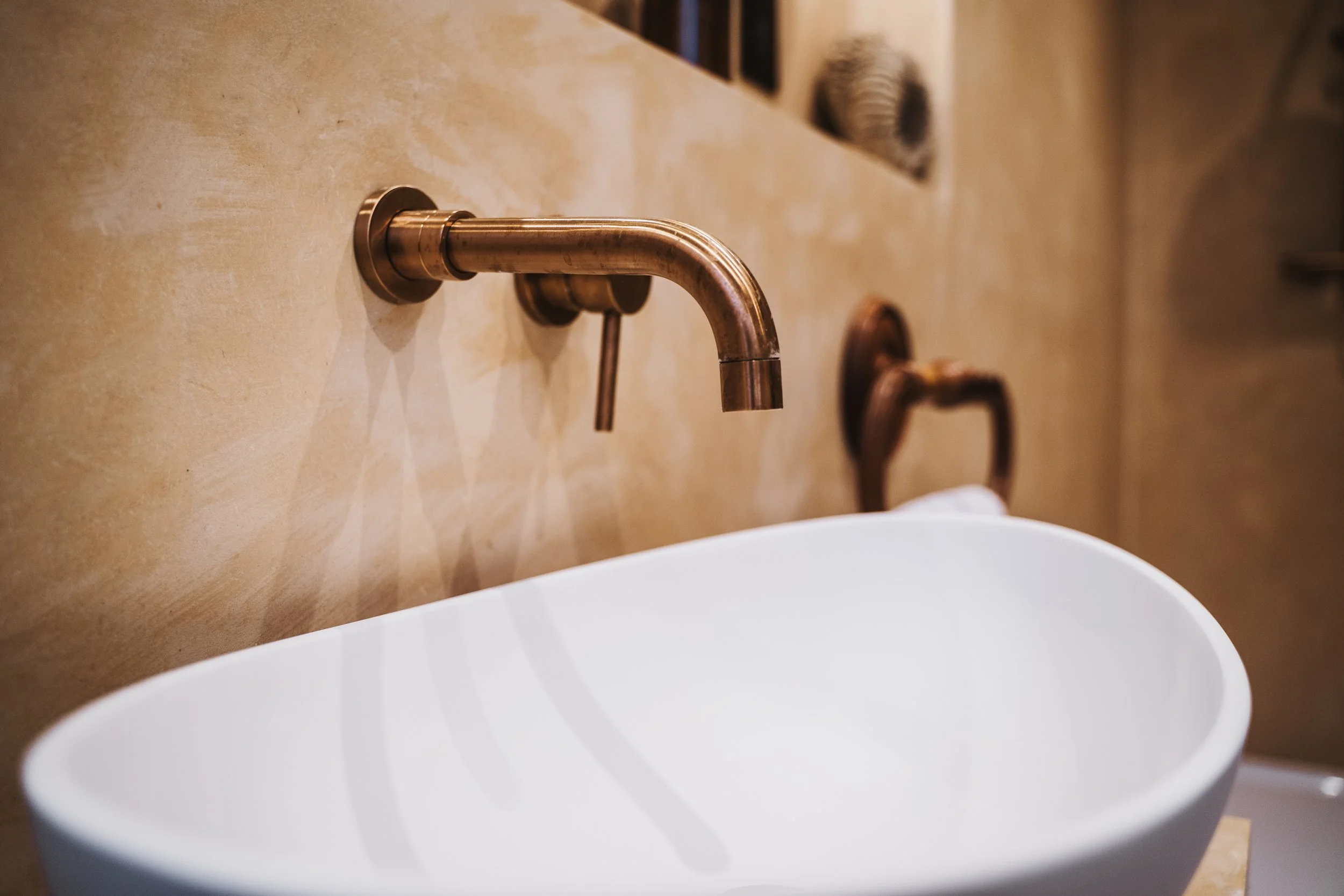 Close-up of a modern, brass-finished wall-mounted faucet above a white ceramic sink in a bathroom with beige textured wall.