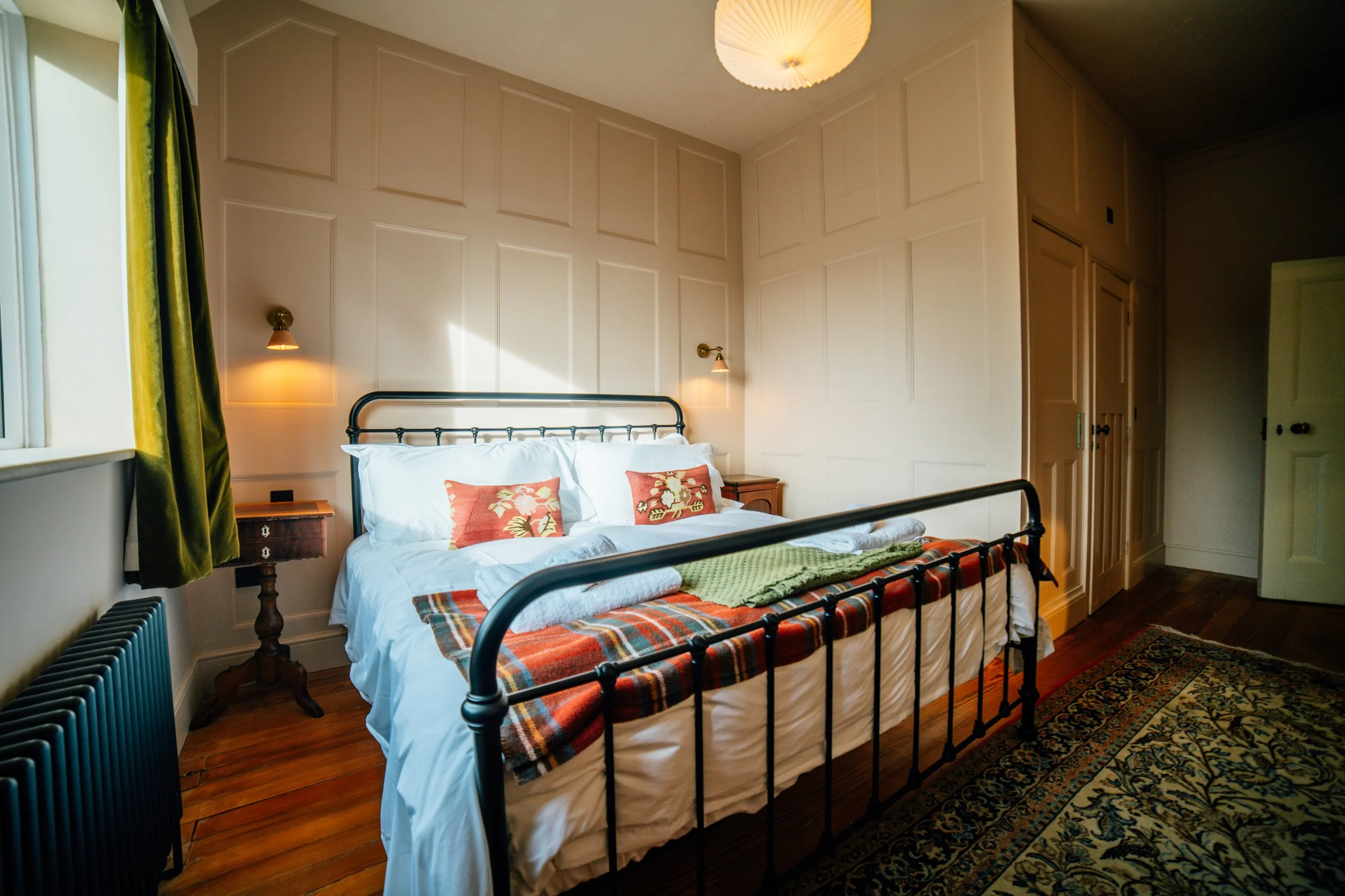 A cozy bedroom with a metal bedframe, white bedding, and colorful pillows. Two wall-mounted lamps provide lighting, and there is a large window with green curtains. A wooden nightstand and a patterned rug complete the room's decor.