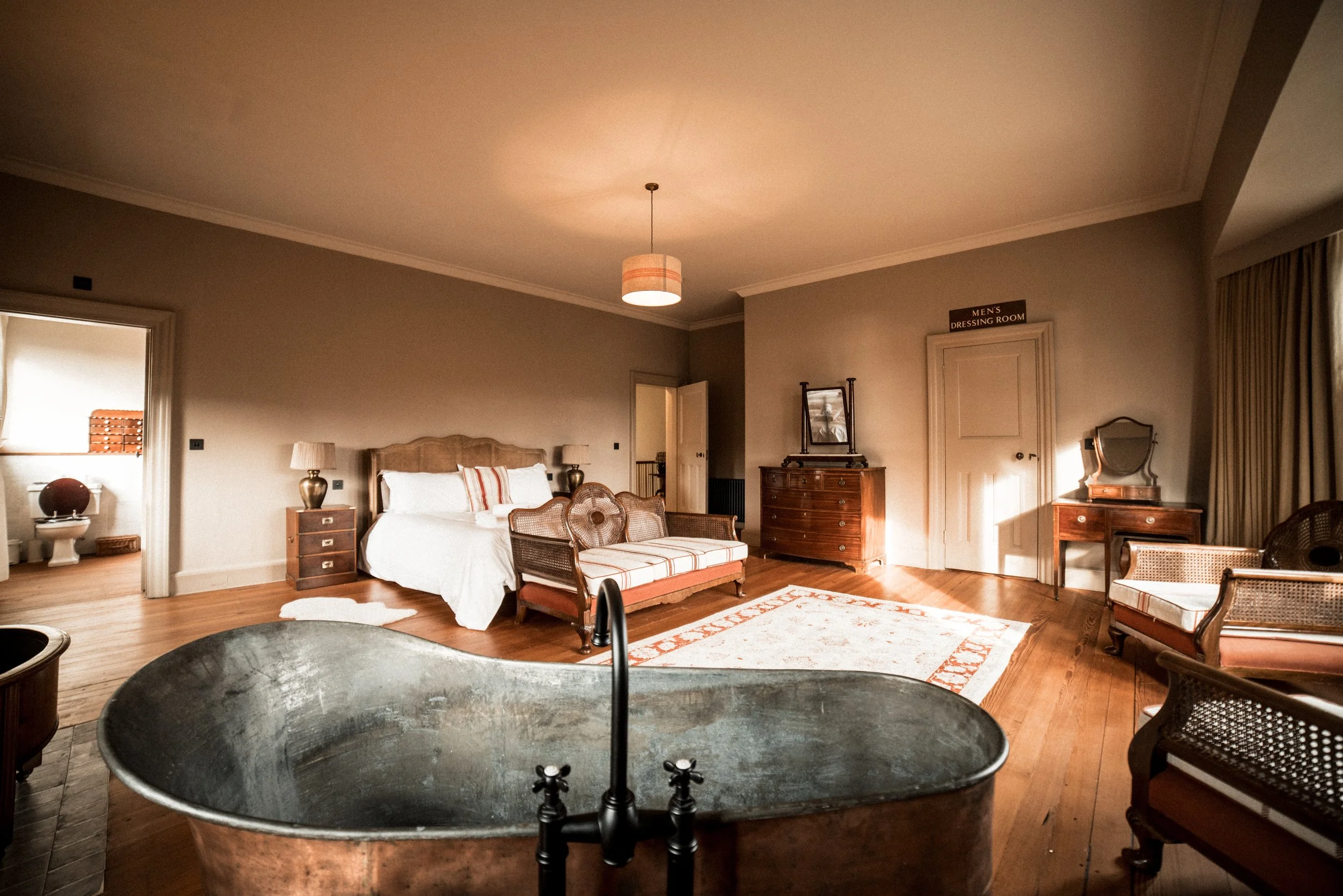 Large vintage bedroom with a clawfoot bathtub in the foreground, a bed with white linens and striped pillows, wooden furniture including a dresser and vanity, a bench, chairs, a rug, and a door labeled 'Men's Dressing Room' in a spacious, warmly lit room.