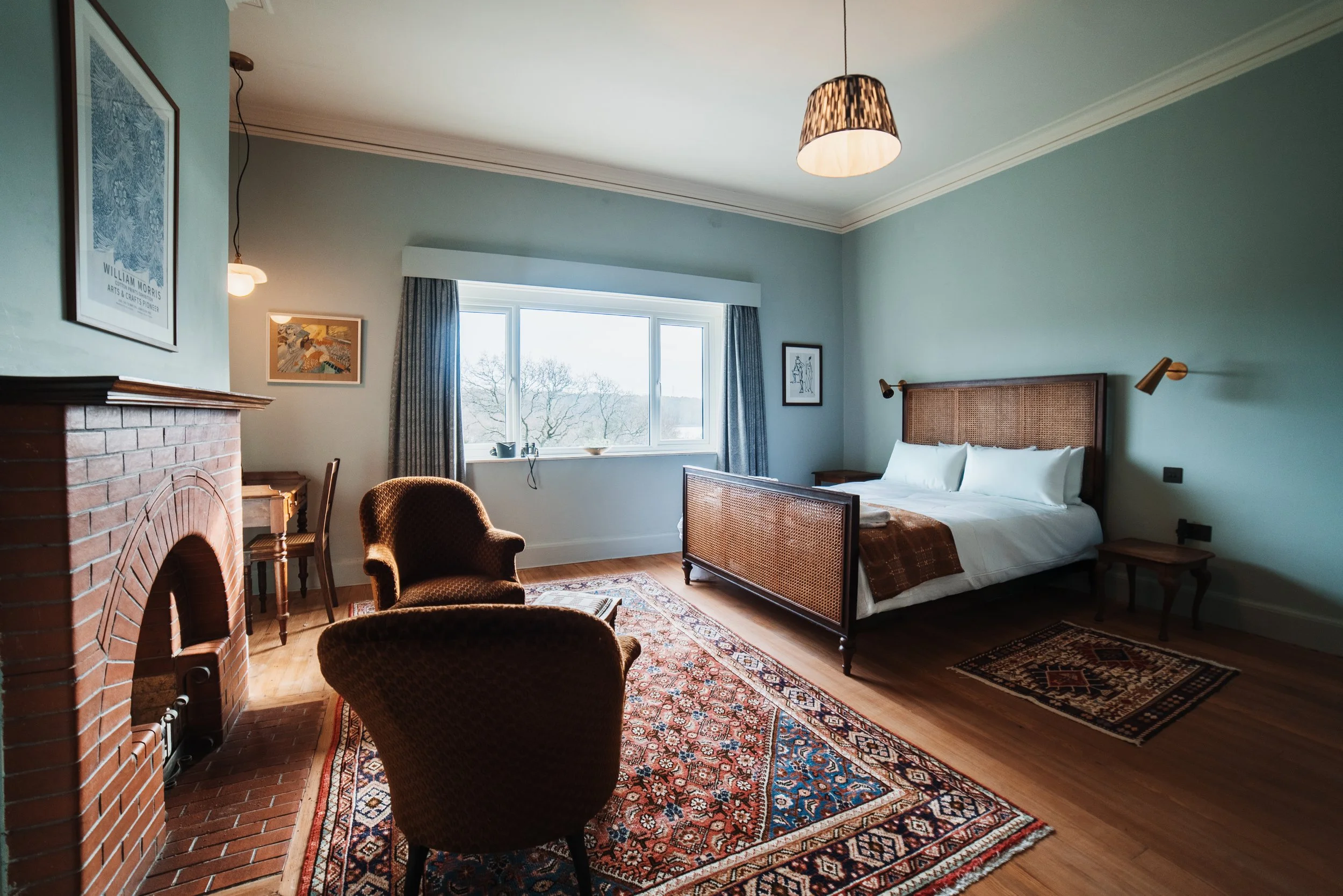 A cozy hotel bedroom with a large window, a bed with a woven headboard, two armchairs, a fireplace, and colorful rugs.