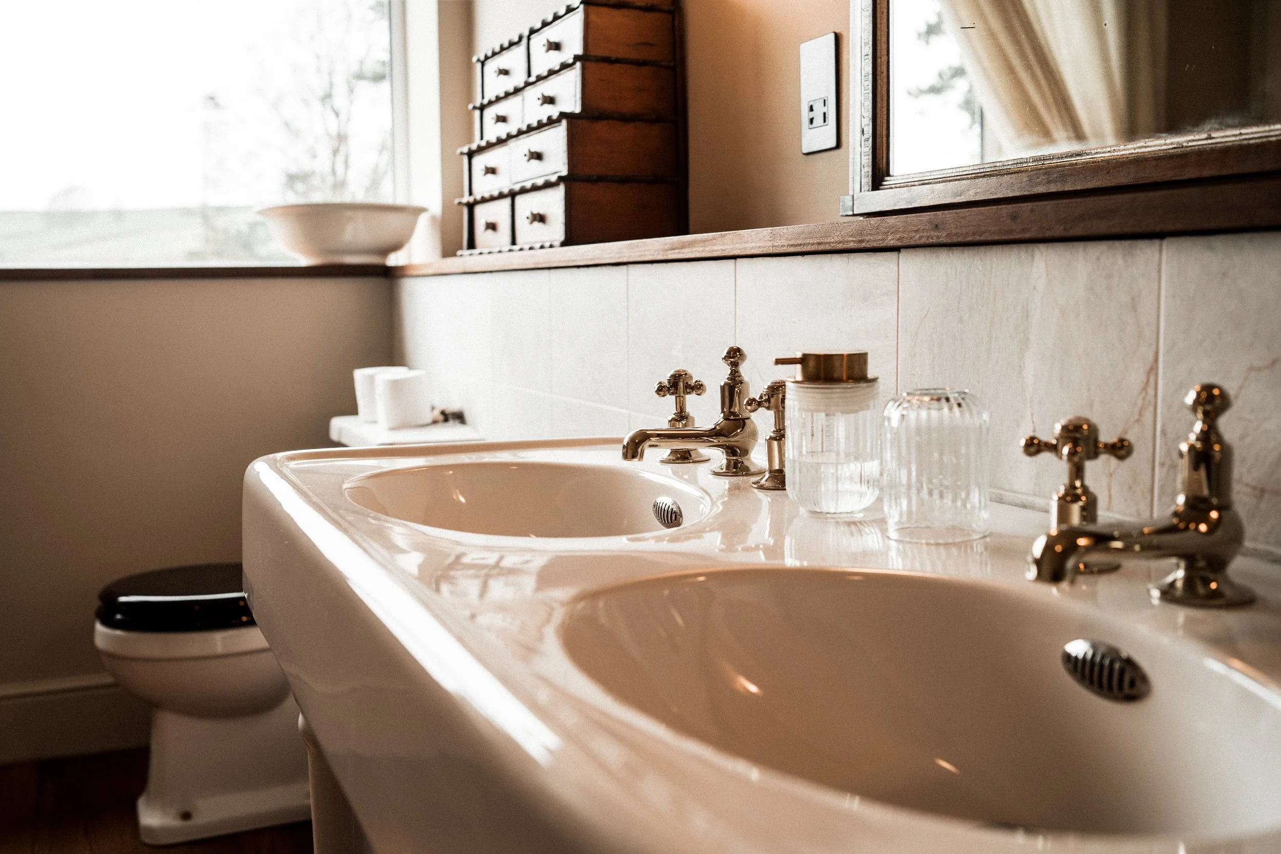 Vintage double bathroom sink with brass faucets, clear glass soap dispenser, and a mirror above, surrounded by beige tiles and wooden accents.
