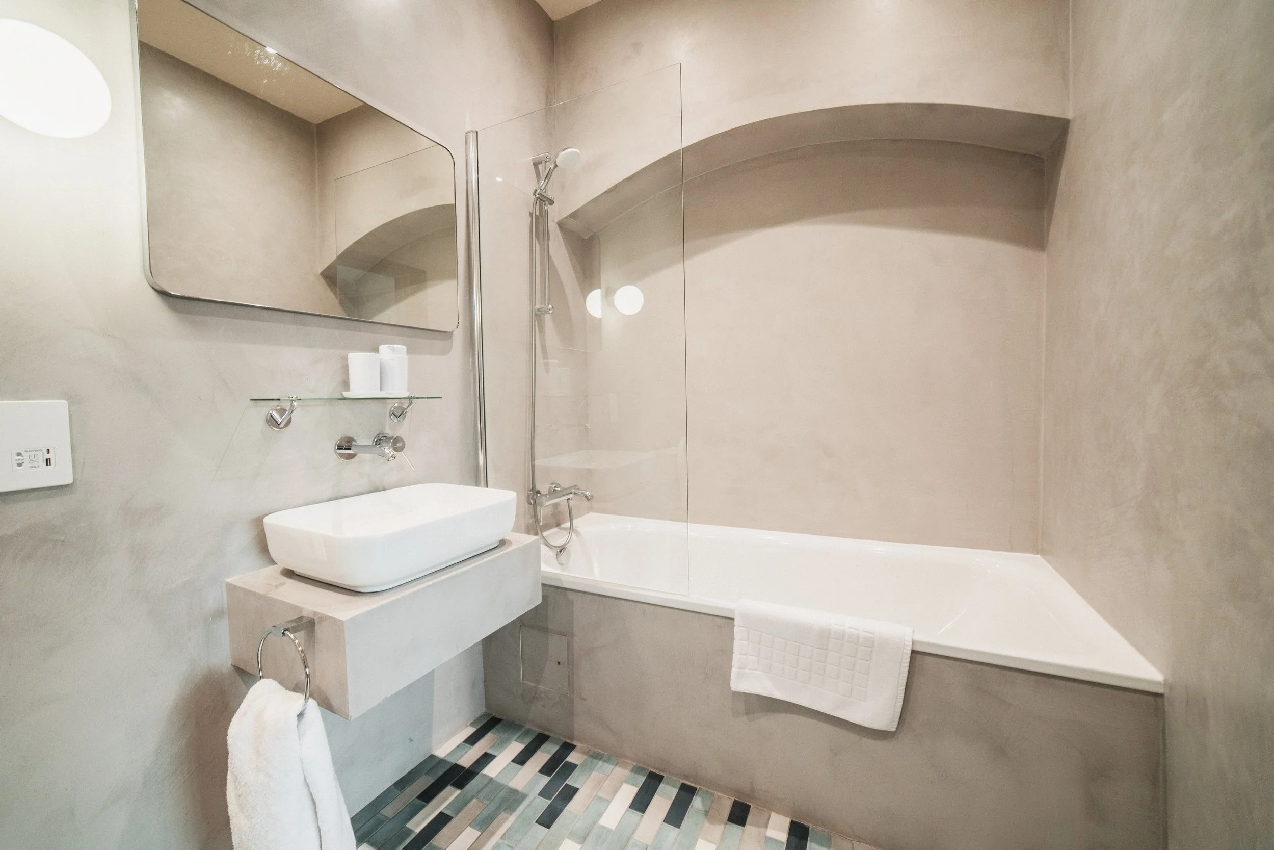 Modern bathroom with beige walls, a bathtub, a wall-mounted sink with a vessel basin, a large wall mirror, and a glass shelf holding toiletries.