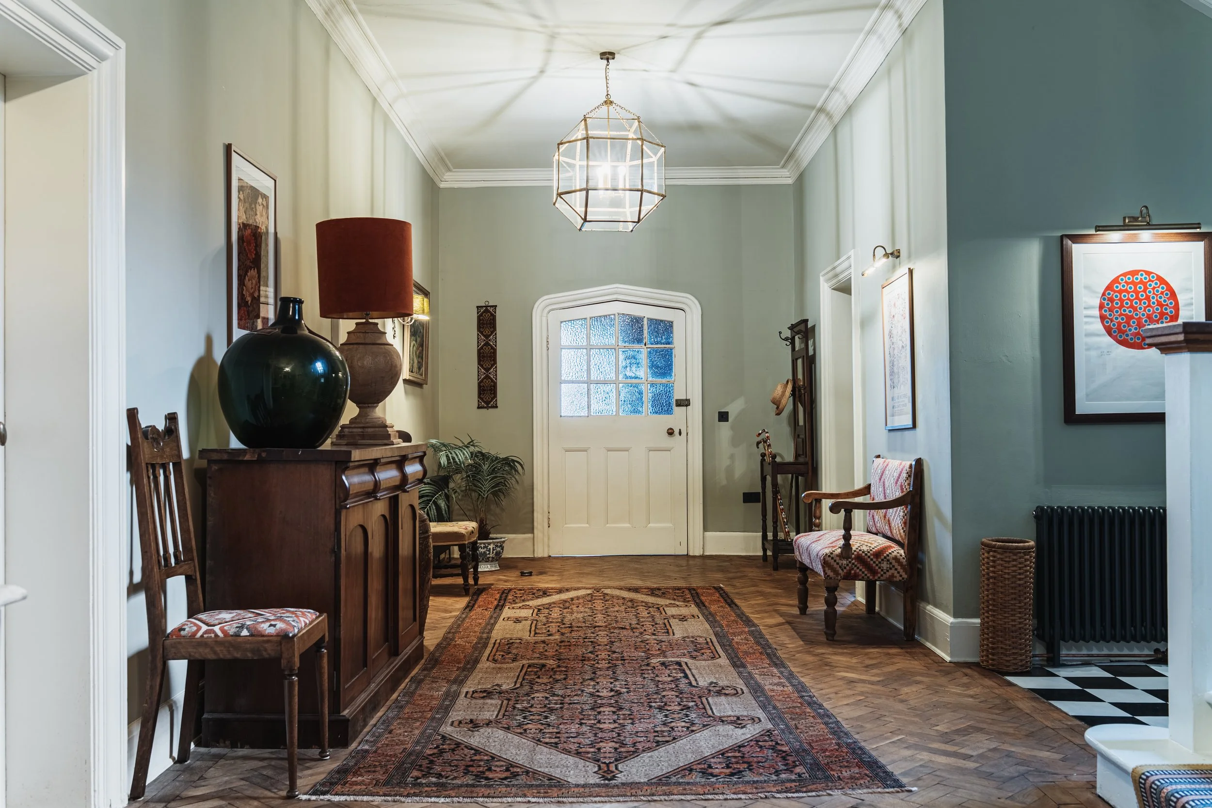 Entrance hallway with vintage and modern decor, including a patterned rug, wooden furniture, framed artwork, a large lamp, and a glass-paneled front door, with hardwood floors and soft-colored walls.