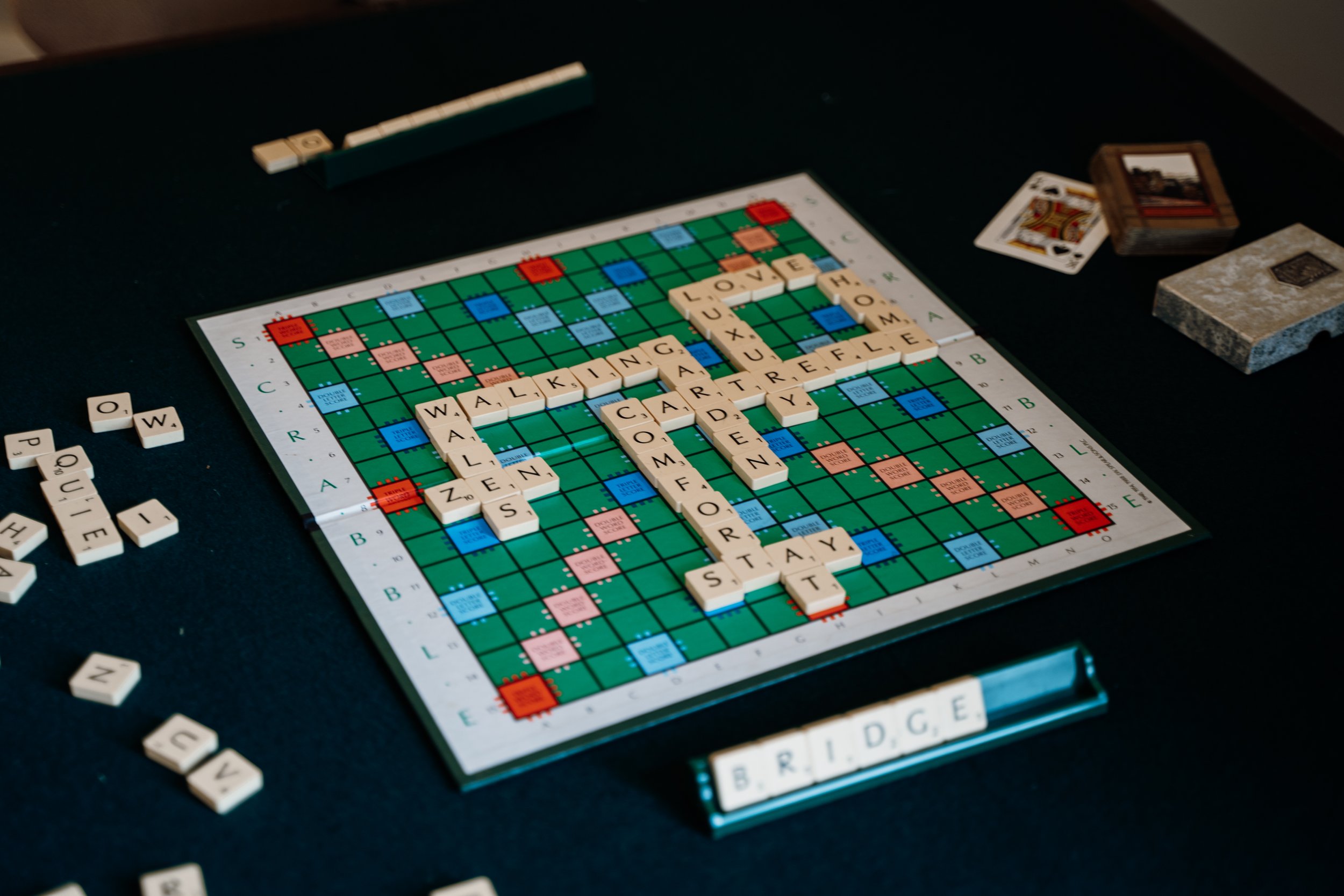 A Scrabble game with words like 'LOVE', 'WALKING', 'CART', and 'HOME' on the board, surrounded by letter tiles and game pieces on a black table.