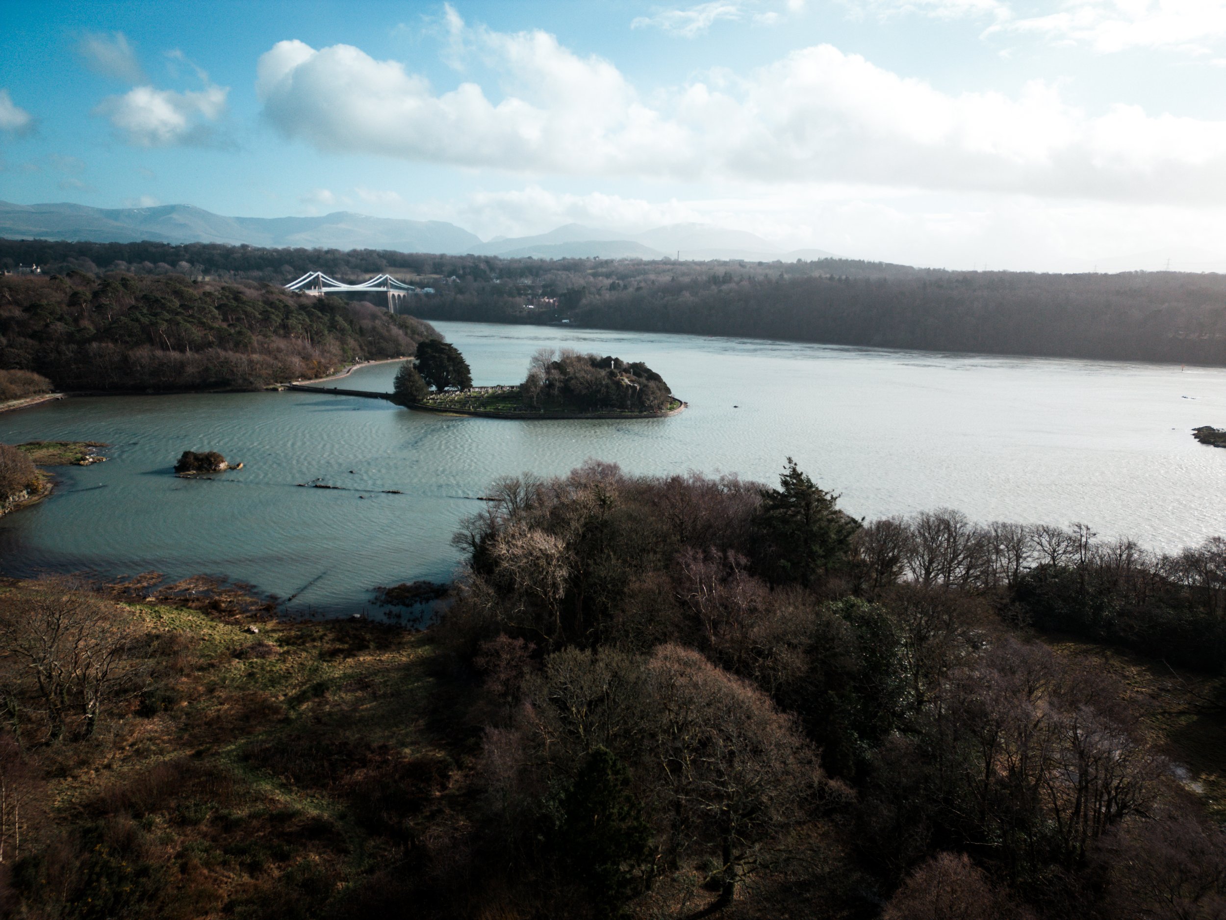 A scenic view of a large body of water with an island in the middle, surrounded by forested land and hills in the background, with a bridge visible in the distance under a partly cloudy sky.