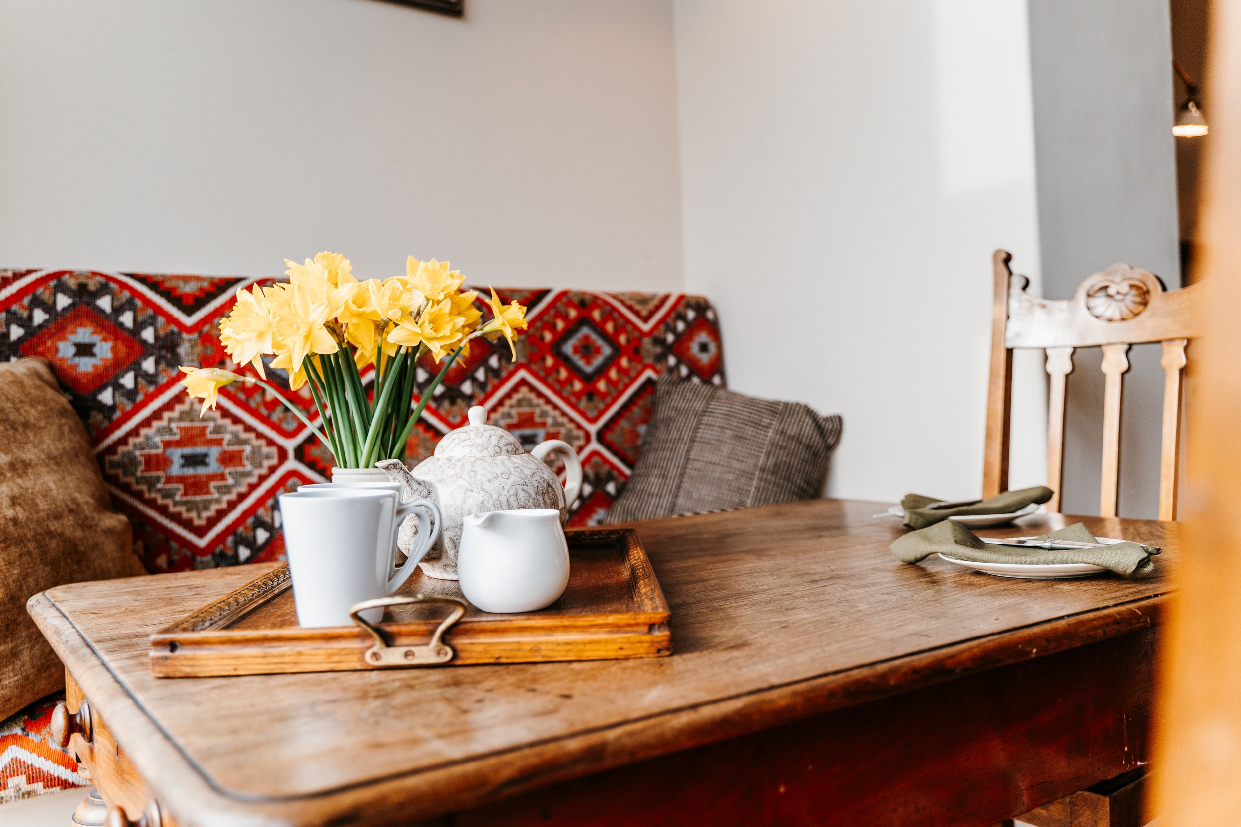 A wooden dining table set with a tray holding a white teapot, mugs, and a small pitcher, with a vase of yellow daffodils, and place settings with green napkins and silverware. In the background, there is a patterned sofa with brown pillows.