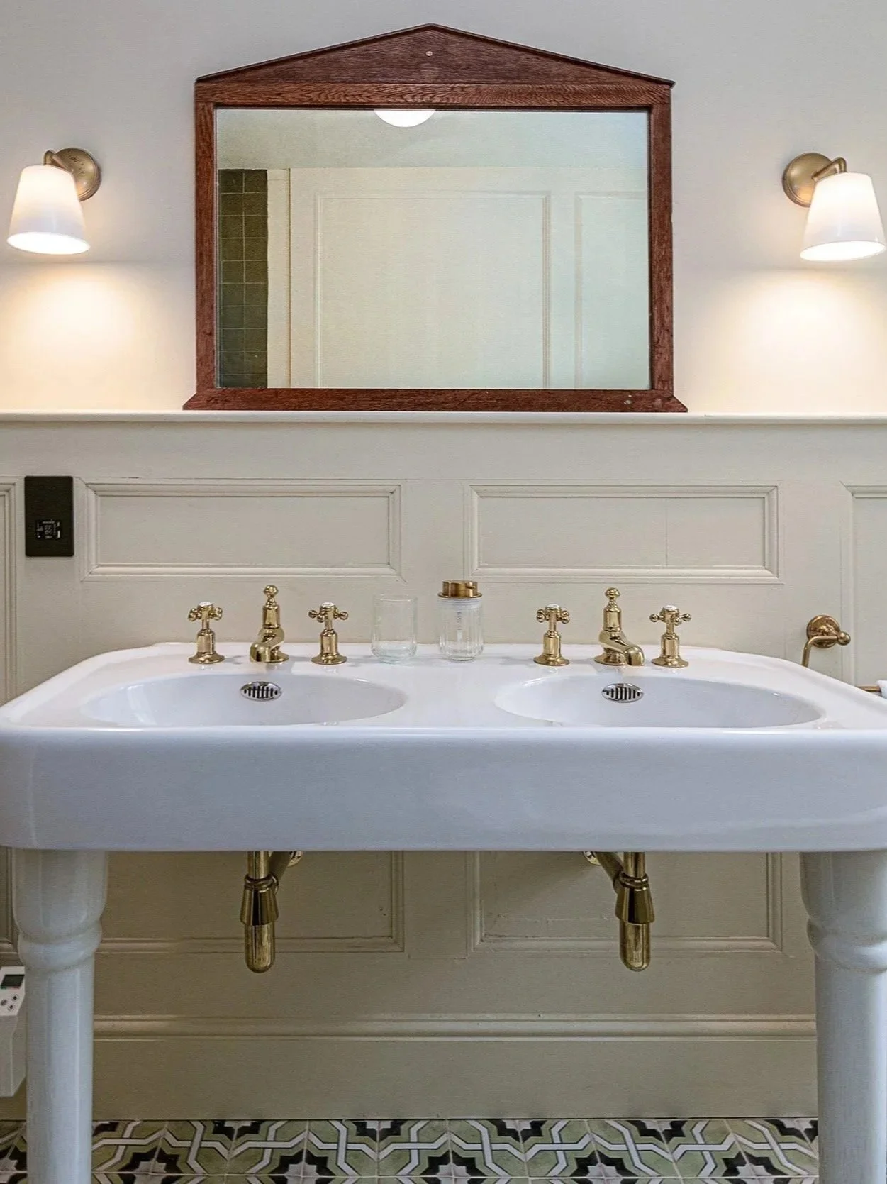 Double sink bathroom vanity with gold fixtures, a large mirror, wall-mounted lights, and patterned floor tiles.