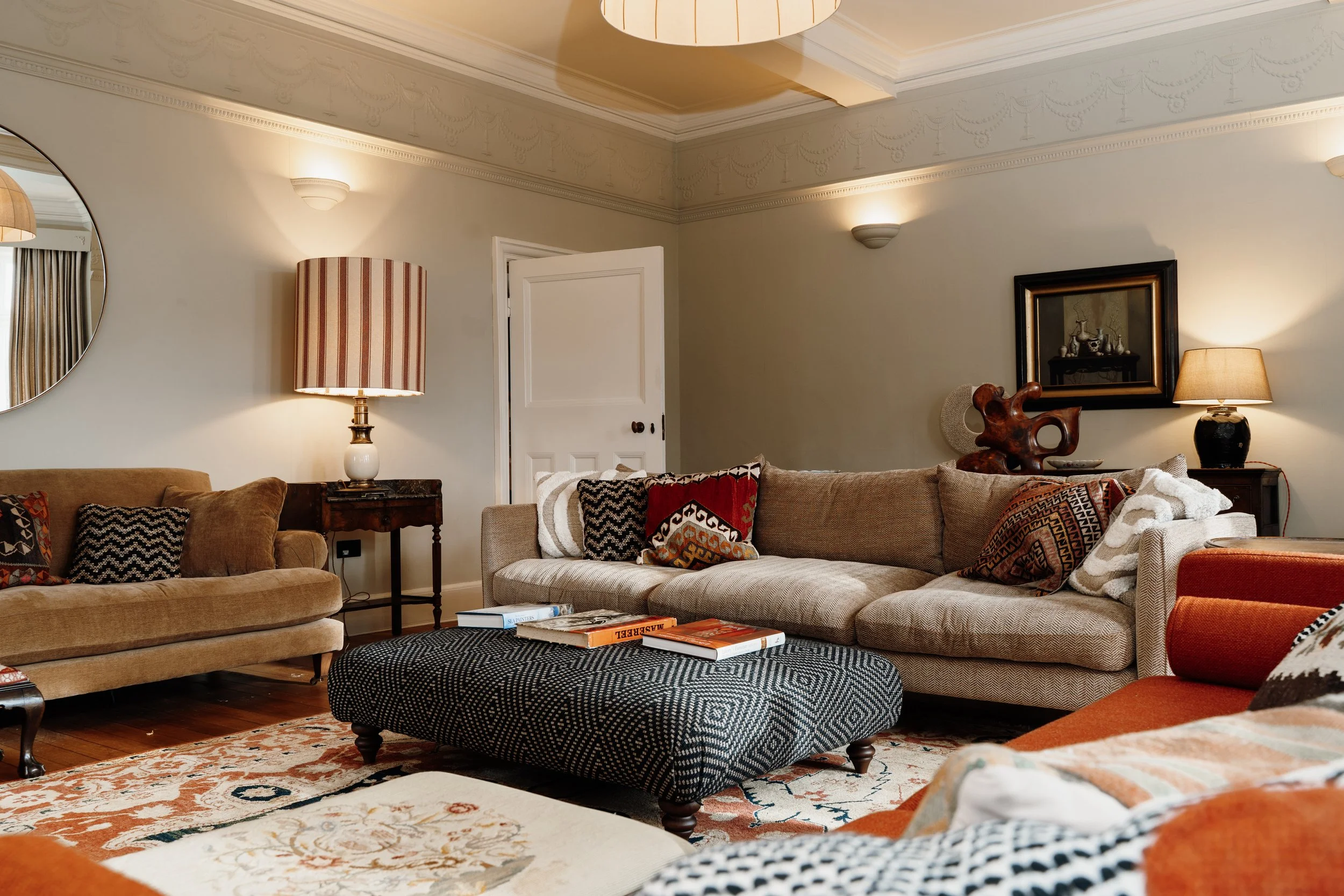 Living room with beige, brown, and orange sofas, a patterned ottoman with books, decorative pillows, a framed picture, lamps, and wall sconces in a cozy, traditional style.