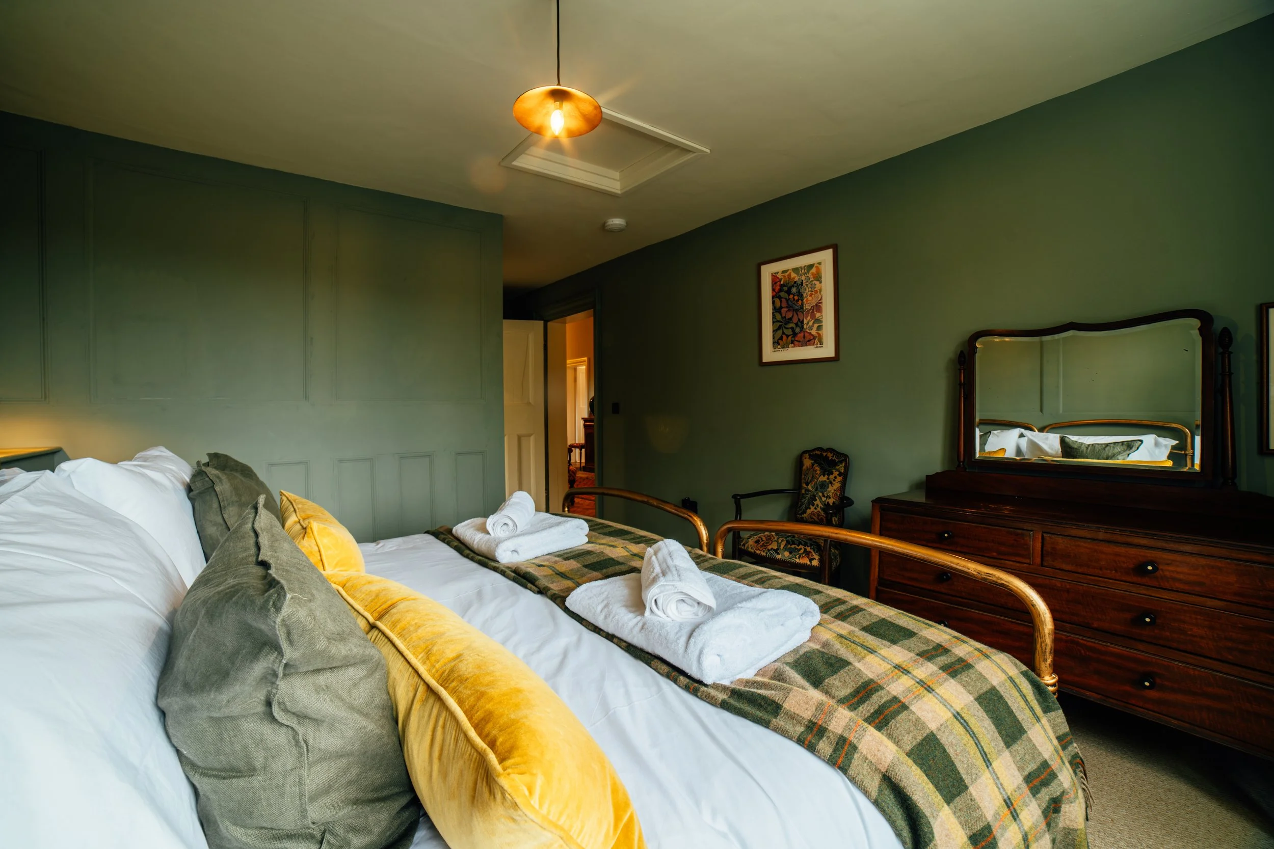 A cozy hotel bedroom with a bed covered in white linens, dark and yellow pillows, and a plaid bed runner. Towels are neatly rolled on the bed. A wooden dresser with a mirror and a chair are on the right. The room has green walls, a ceiling light fixture, and framed artwork on the wall. An open door shows a glimpse into another room.