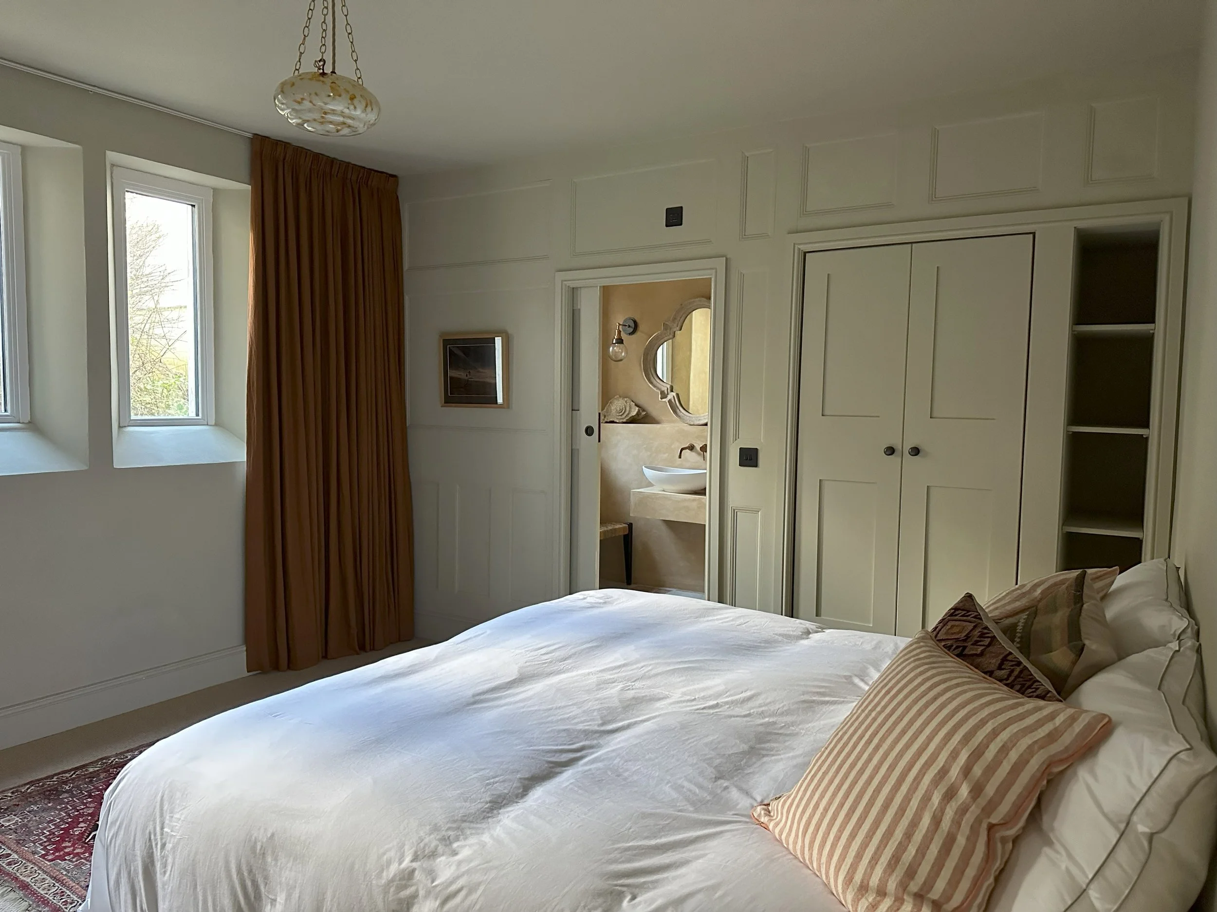 A bedroom with a white bed, pillows, windows, a curtain, and an open door revealing a bathroom with a sink and mirror.