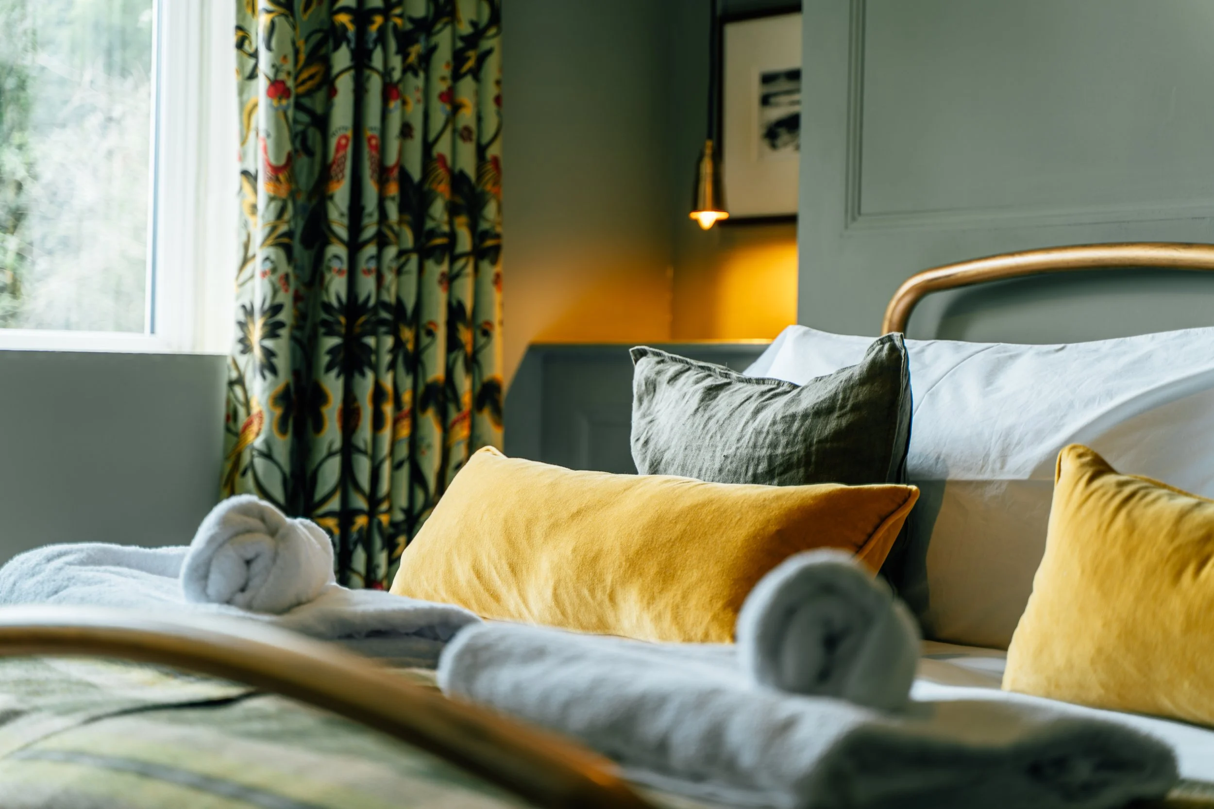 A neatly made bed with decorative pillows in various colors, including yellow, dark green, and white. Towels are rolled at the foot of the bed. A window with floral patterned curtains is visible to the left, allowing natural light to brighten the room. A warm light is on beside an artwork on the wall.