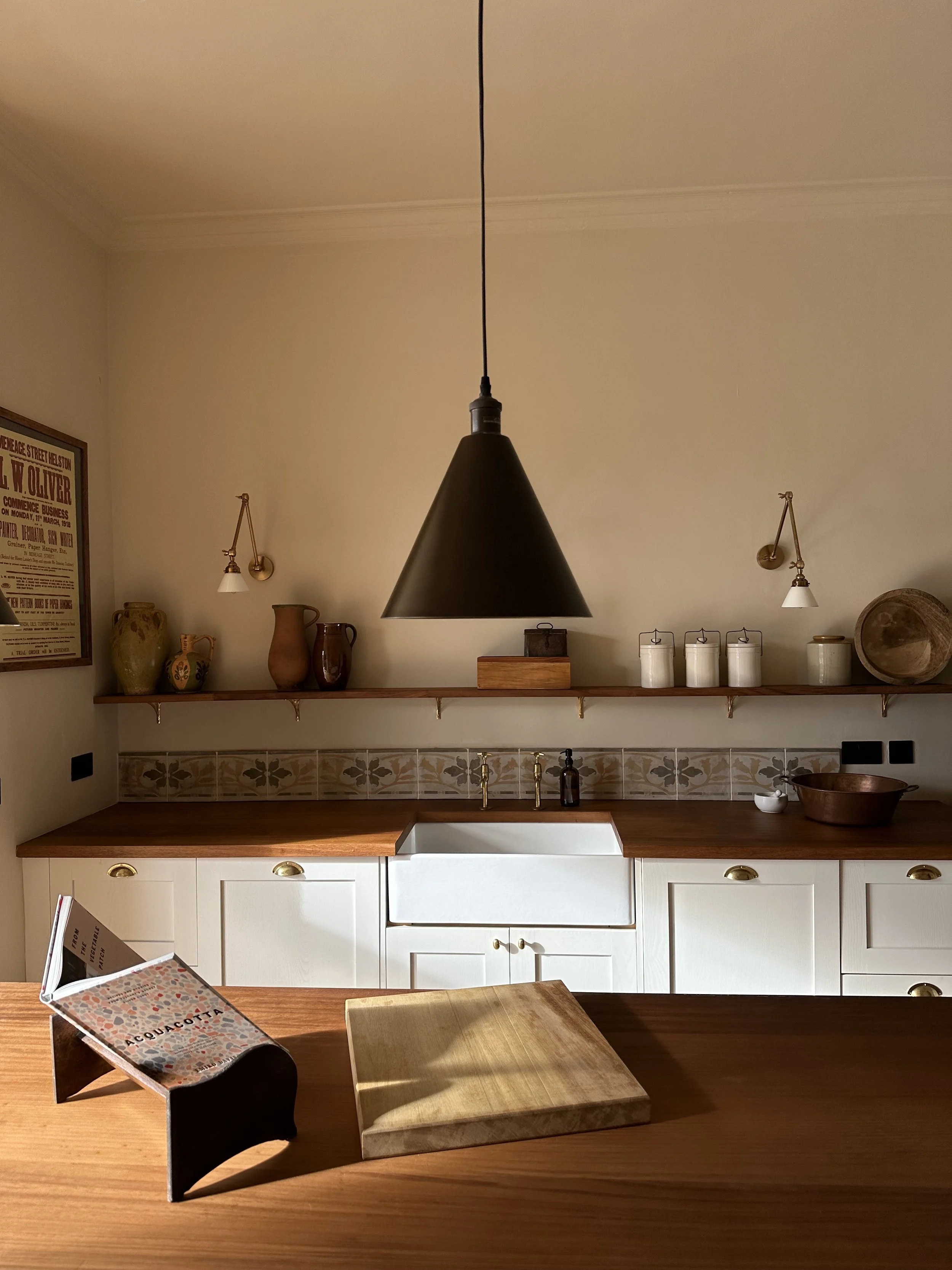 A modern kitchen with a wooden countertop, white cabinets, decorative vases, a wooden shelf with jars and bowls, a black pendant light, and a vintage advertisement poster hanging on the wall.