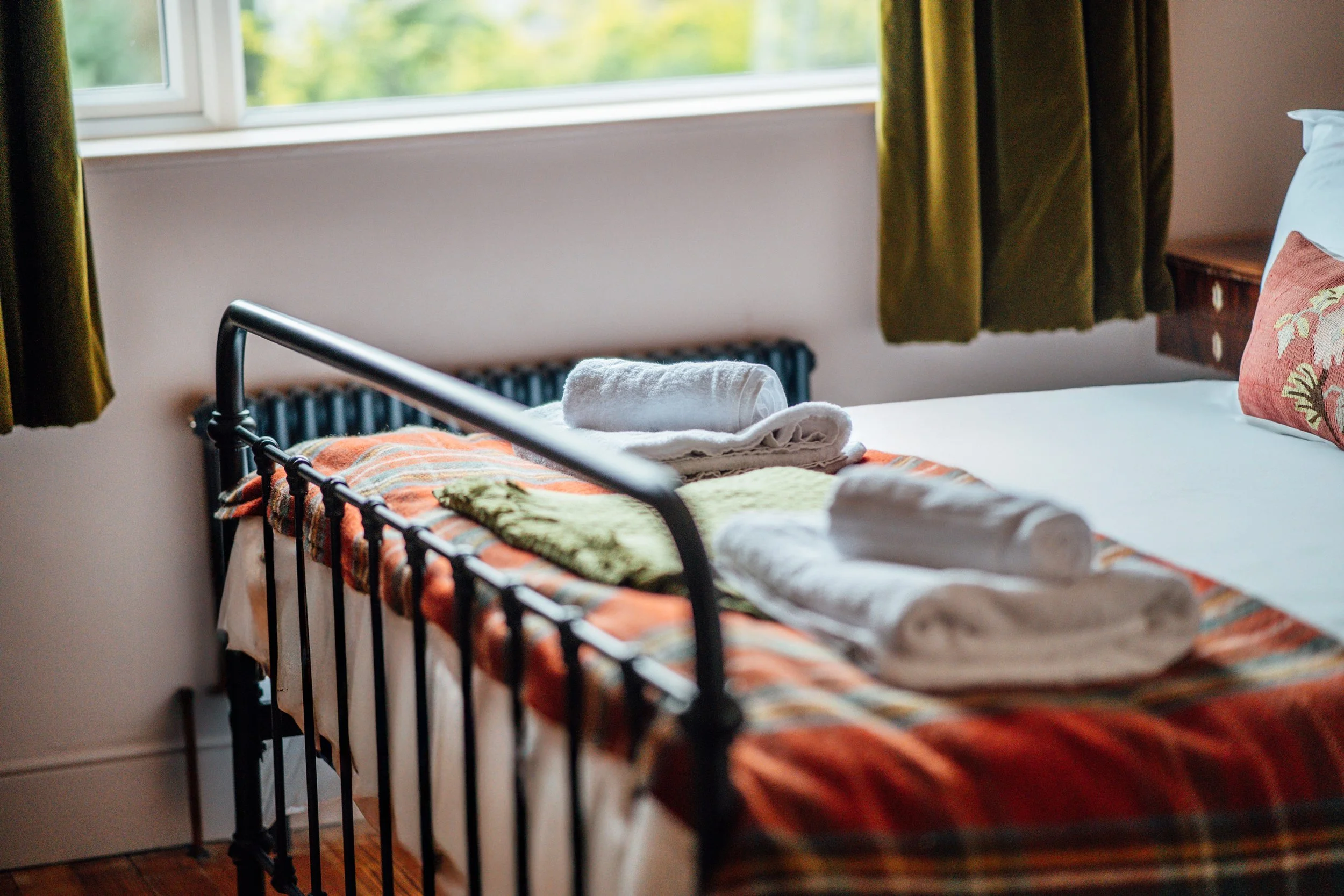 A neatly made bed with rolled white towels and green washcloths on top, a plaid blanket, and a decorative pillow, with green curtains and a window showing a blurred outdoor view.
