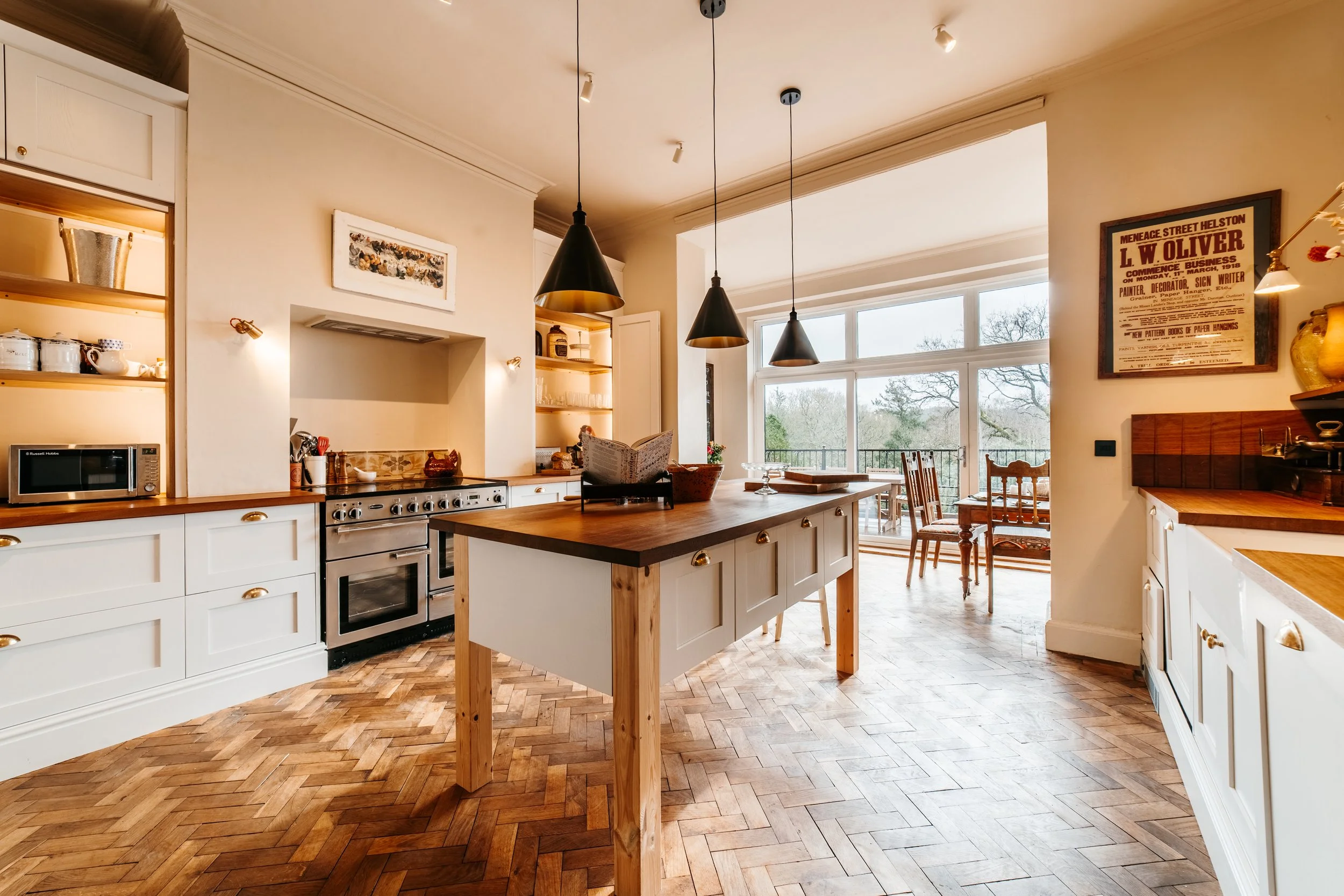Bright kitchen with white cabinets, wooden countertops, black and gold pendant lights, and large windows overlooking trees.
