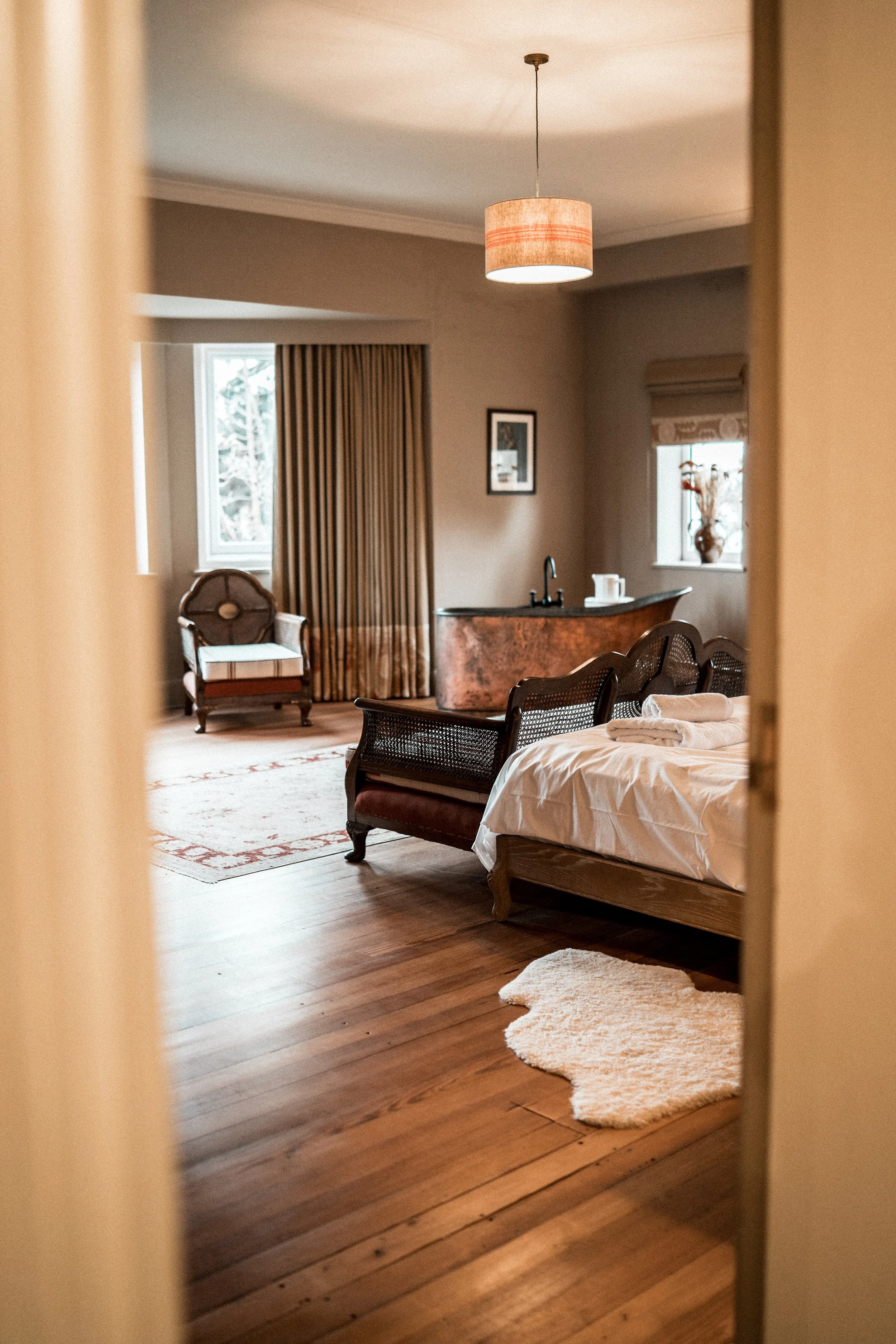 View of a cozy bedroom with wooden floors, a bed with linen, a fluffy rug, a warm pendant light, armchair, window with curtains, and a small vanity or counter with a vase.