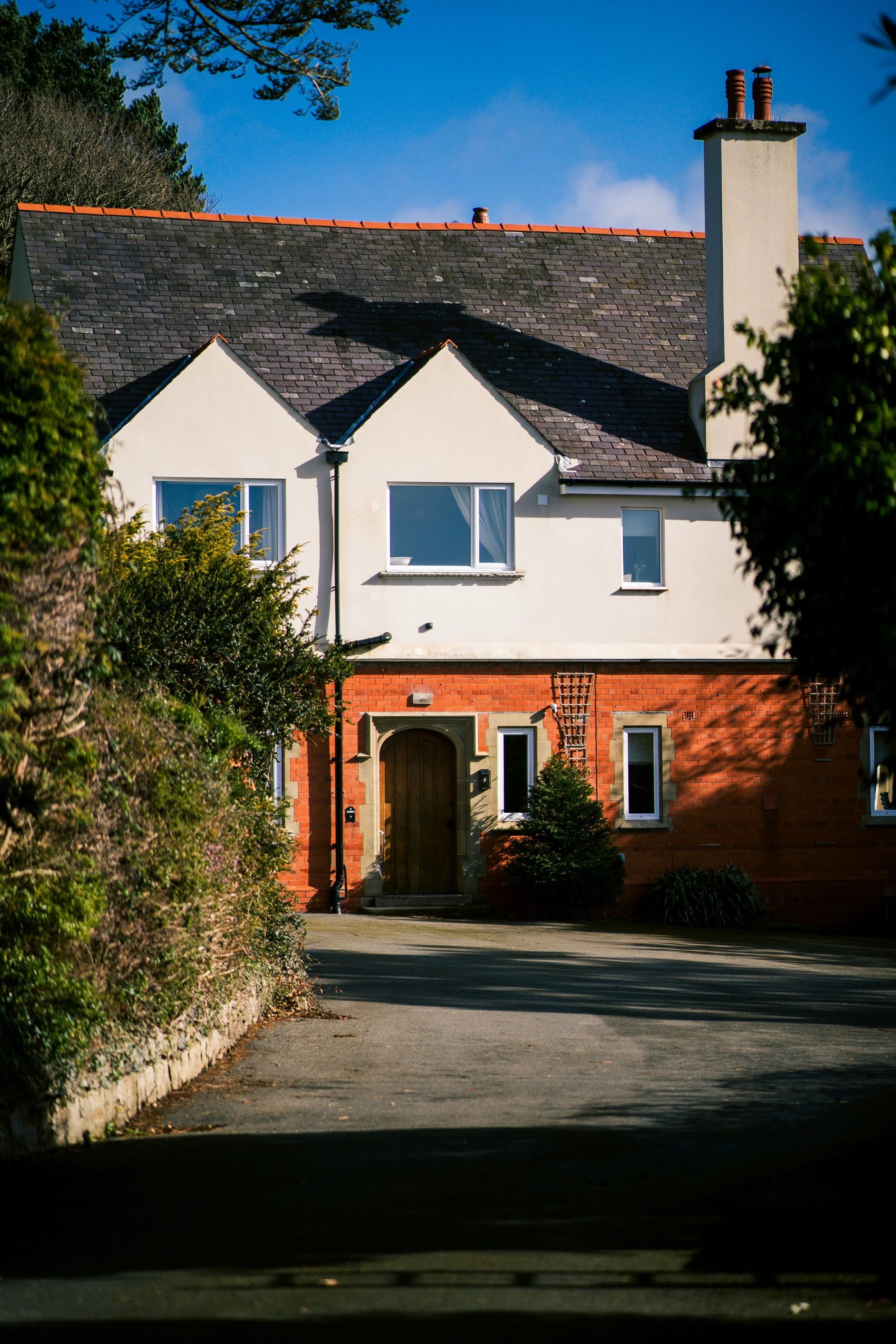 A two-story house with a red brick lower level and white upper level, with multiple windows and a wooden front door, surrounded by greenery, under a partly cloudy blue sky.