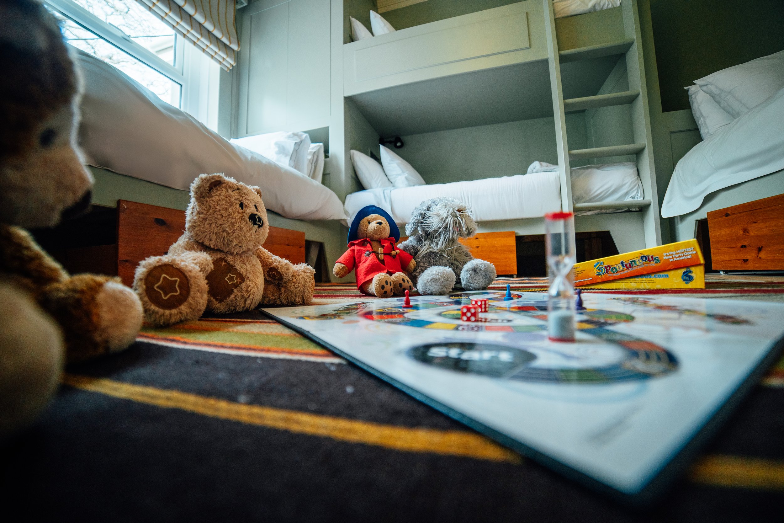 Toys including teddy bears, a stuffed dog, and a toy doll arranged around a board game on a colorful striped carpet in a bedroom with bunk beds in the background.