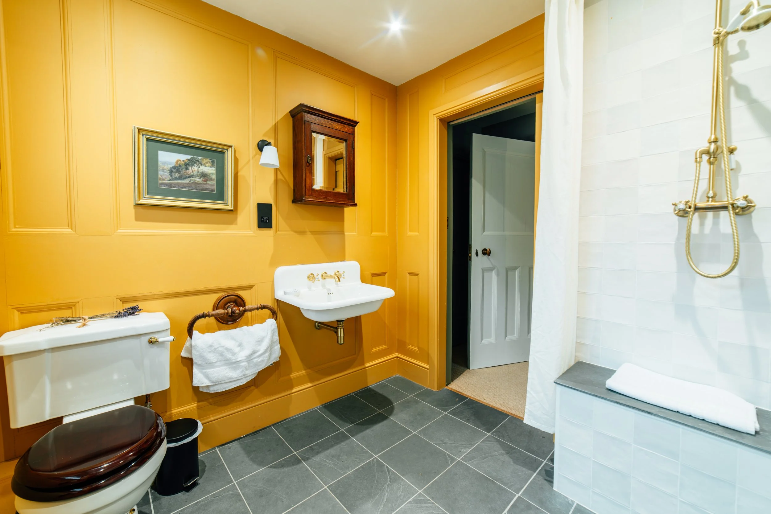 A bathroom with yellow paneled walls, a white toilet with a dark wood seat, a small white sink, and a shower area with a white curtain and a gold-colored showerhead. Includes a framed landscape painting, a wooden medicine cabinet, and a towel rack with a white towel.