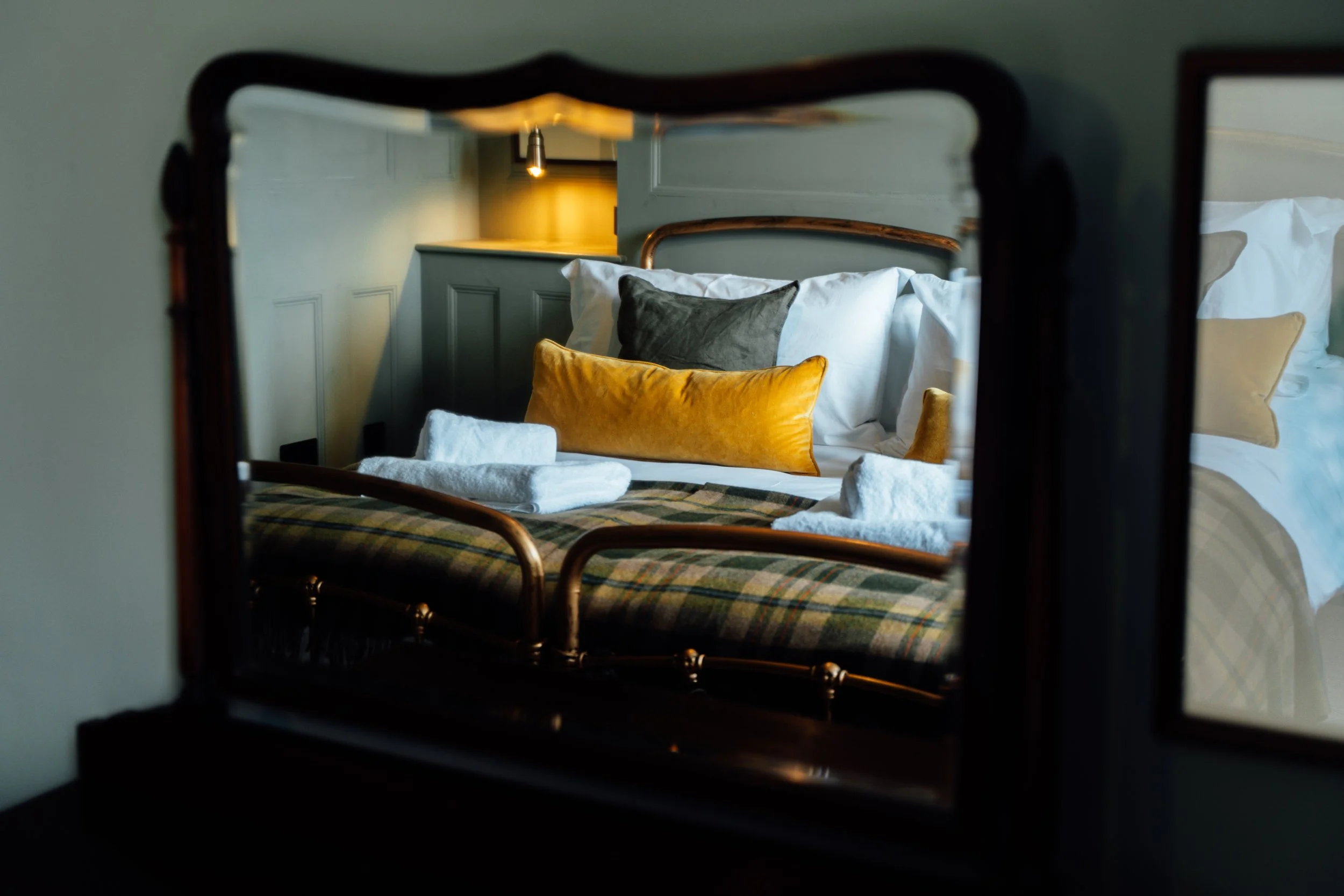 A bed with white linens, decorated with black, gold, and beige pillows, seen through a mirror with a dark wood frame.