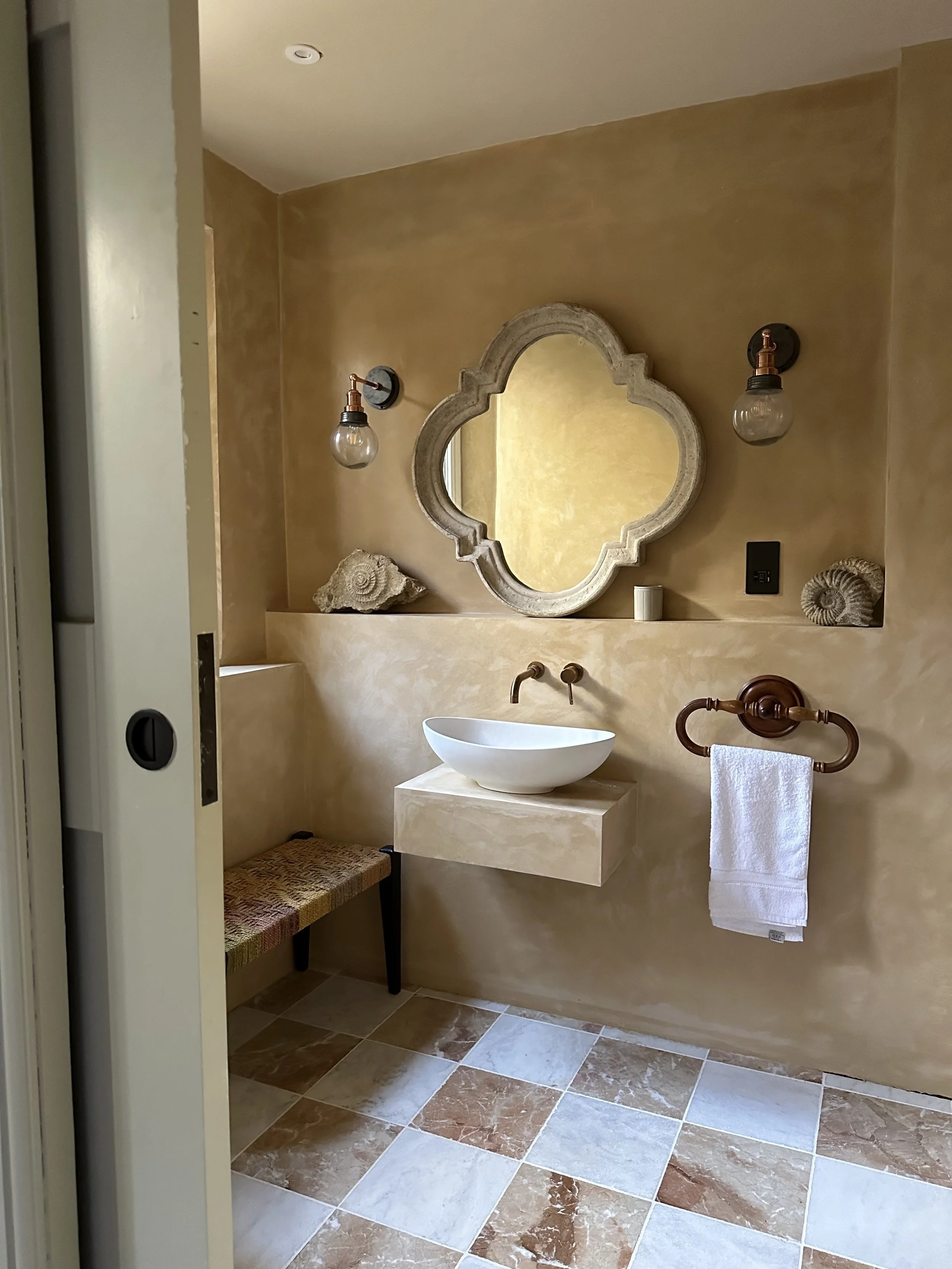 A bathroom with beige textured walls, a floating beige sink with a round vessel basin, and a decorative mirror with a quatrefoil shape. There are two wall-mounted light fixtures with exposed bulbs, seashell decorations on the ledge, a small woven bench, and a white hand towel hanging on a vintage-style bronze towel ring.