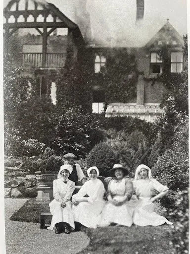 Four women dressed in vintage clothing and wide-brimmed hats sitting on a bench in a garden with a house in the background.